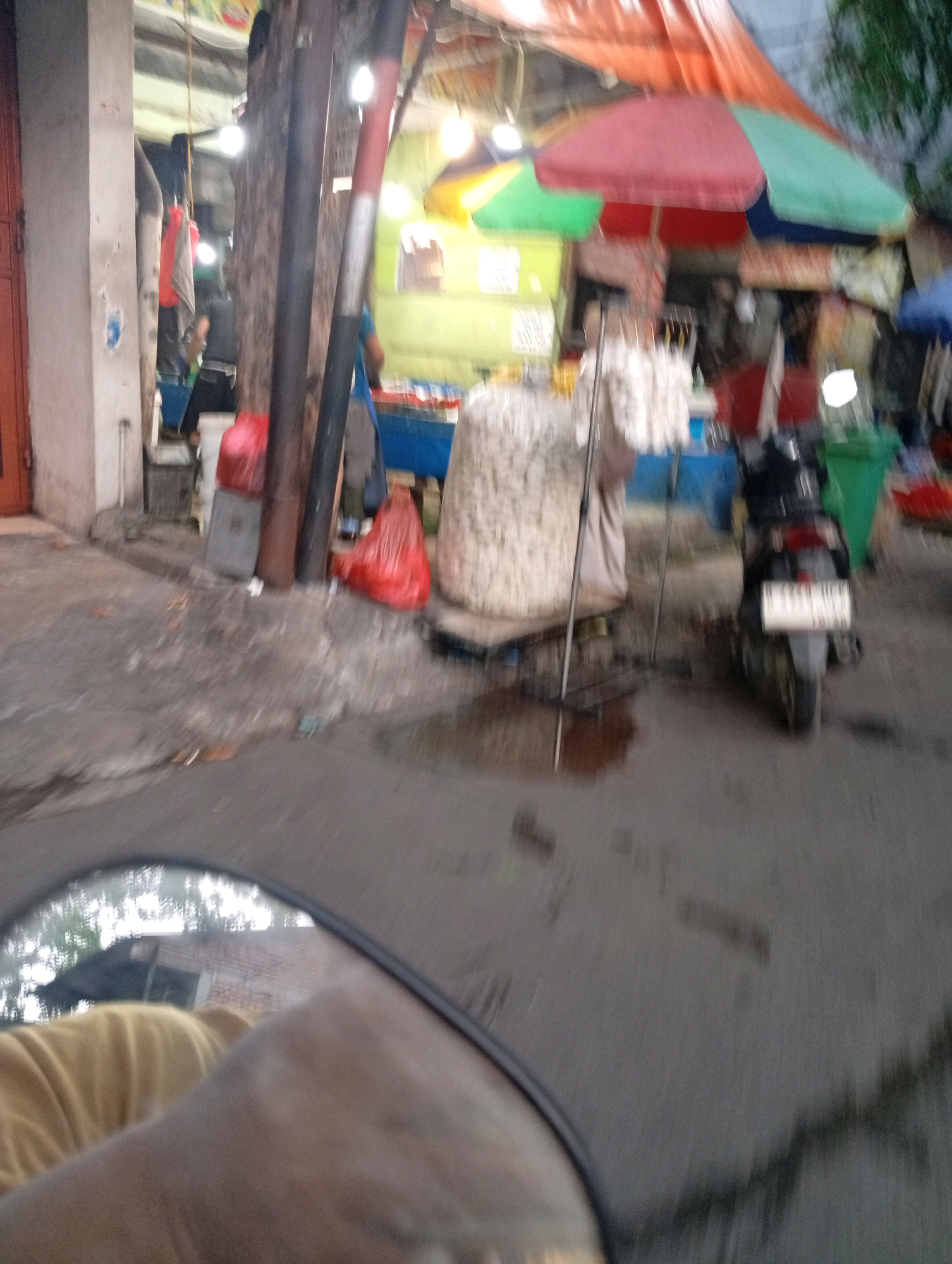 The image depicts a street scene in North Jakarta, Indonesia, likely a market area. In the foreground, a blurry reflection in a mirror shows a person wearing a yellow shirt. Behind the mirror, the scene unfolds with a paved road that appears wet, suggesting recent rain. Several stalls are visible, covered by colorful tarps, with some items displayed.
To the left, a building with a white facade and a red door stands. A vertical pole, possibly a lamppost or part of a stall structure, is prominent in the mid-ground. Red plastic bags are seen on the ground near the pole. A large, light-colored, cylindrical object, possibly stacked goods or a large container, is positioned on a stand.
A black motorcycle is parked on the right side of the road, facing away from the viewer, with a license plate that is not clearly legible. Further back, green and orange umbrellas are visible over a stall. Other blurry figures and objects suggest activity in the market. The overall atmosphere is that of a bustling, possibly slightly disorganized, street market in an urban environment.