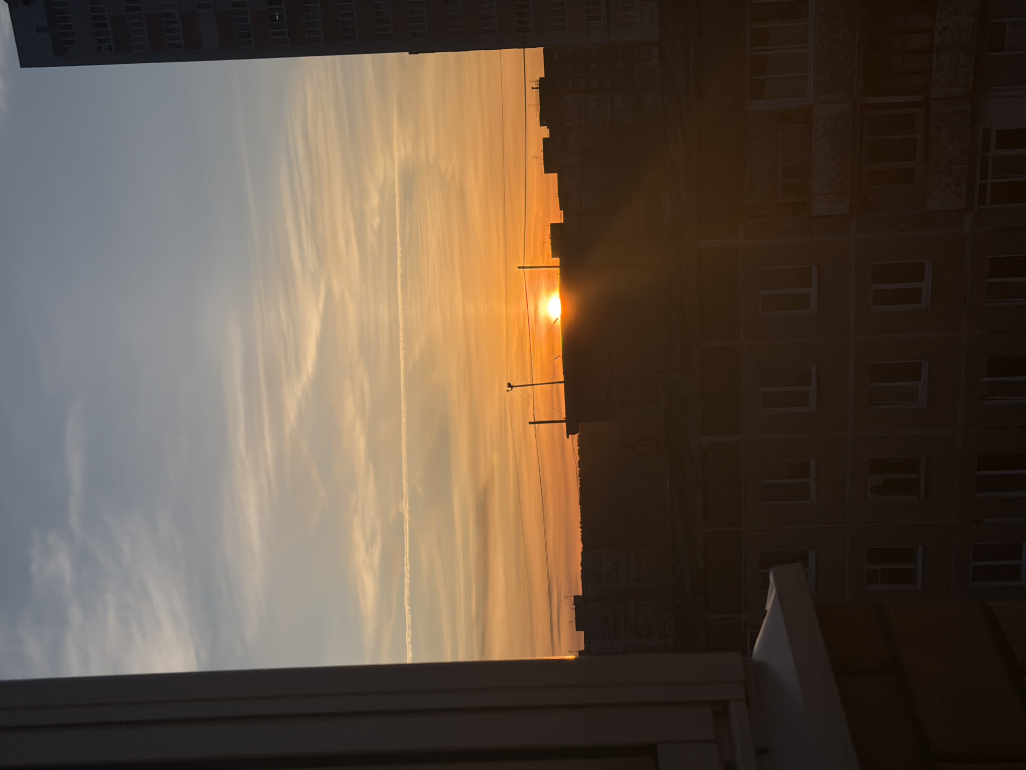 An urban sunset is captured from an interior vantage point, looking out over a city, likely Perm, Russia. The image is oriented vertically but depicts a horizontal panorama. The sky dominates the upper and left portions of the scene, transitioning from a soft, light blue-grey at the top left to a warm, vibrant orange and yellow hue towards the horizon line, where the sun is visible. Wispy, elongated clouds stretch across the sky, with a distinct, straight contrail prominent in the upper left section.
The bright, fiery sun, positioned low on the horizon slightly right of center, emits distinct rays of light, illuminating the clouds and creating a radiant glow. Below the sun, the silhouettes of numerous urban buildings form a dark, jagged skyline against the colorful sky. Several utility poles with overhead wires are also sharply silhouetted near the sun.
In the foreground, a dark, partial structure, possibly a window frame or balcony railing, is visible in the bottom left corner. On the right side, a tall, multi-story building, also largely in shadow, occupies the mid-ground, showing faint details of its numerous rectangular windows. Another building is silhouetted along the top edge of the frame. The overall atmosphere is serene and contemplative, characteristic of either sunrise or sunset, with the strong golden and orange tones suggesting a sunset. There is no visible text in the image.