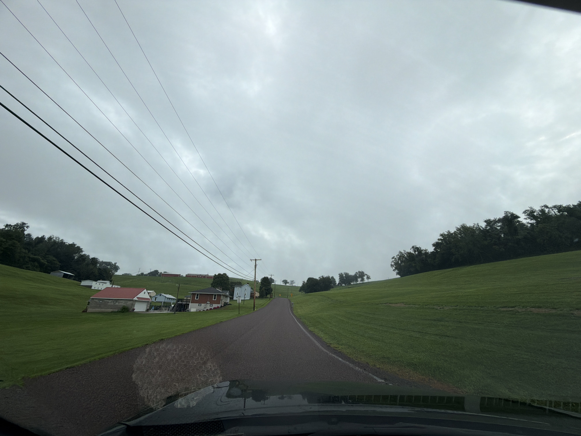 The image is a point-of-view shot from inside a vehicle driving on a wet, asphalt road in Fallowfield Township, USA. The road winds uphill through a rural landscape under an overcast sky. To the left, a grassy slope is dotted with several houses, including a prominent one with a red roof and a detached garage with a red roof. Further up the hill, more houses and buildings are visible. A line of utility poles and power lines runs parallel to the road on the left. To the right, a large, continuous grassy hill slopes upwards, meeting a dense line of trees at the crest. The overall impression is of a quiet, possibly rainy day in a country setting.
