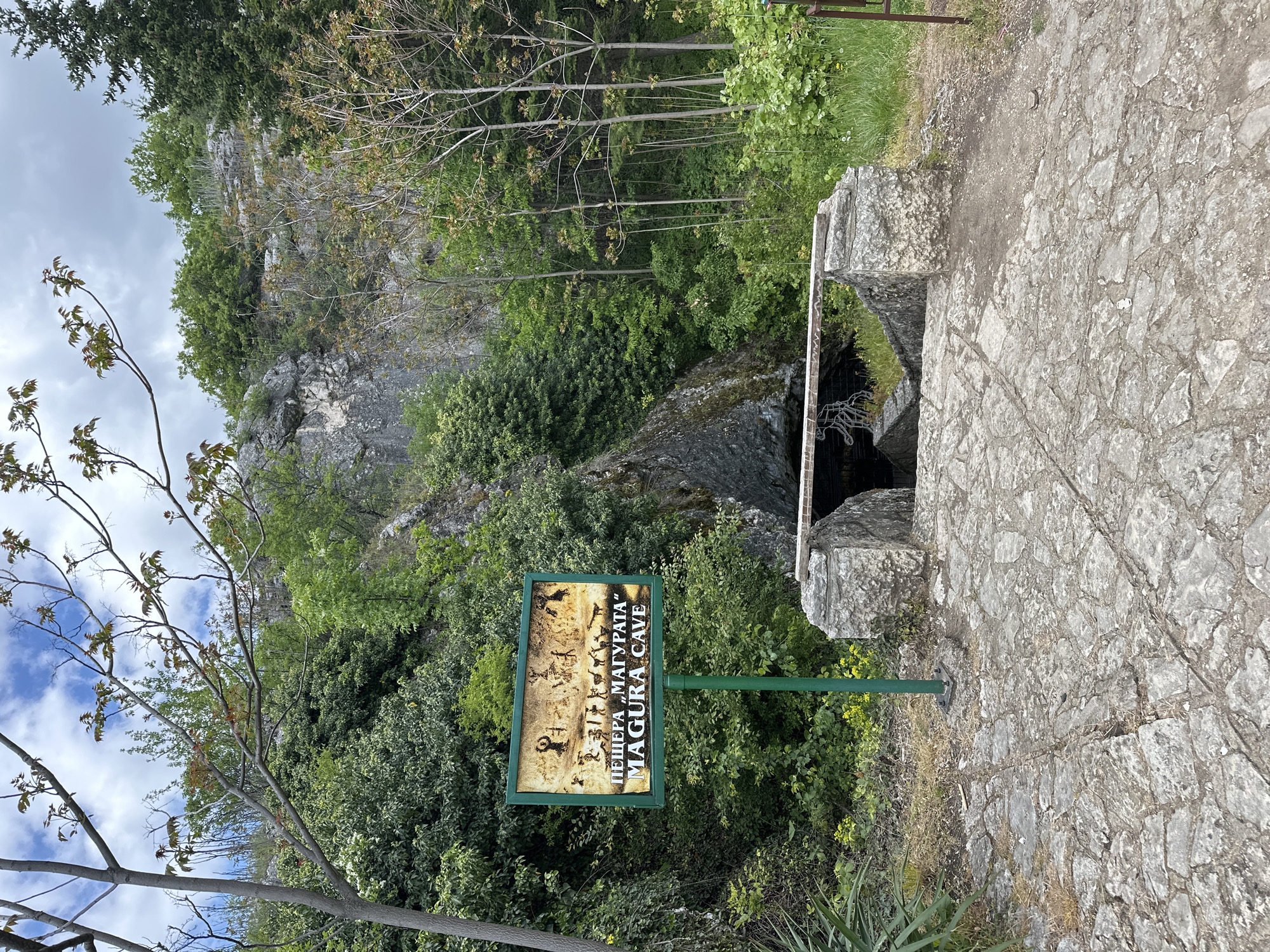 The image shows the entrance to Magura Cave, near Raianovtsi, Bulgaria. The entrance is framed by stone blocks and a metal grate. A sign for "Magura Cave" is positioned nearby. The area is vegetated with trees and bushes. The background features a rocky cliff. The foreground is a stone path. The sky is partially cloudy.