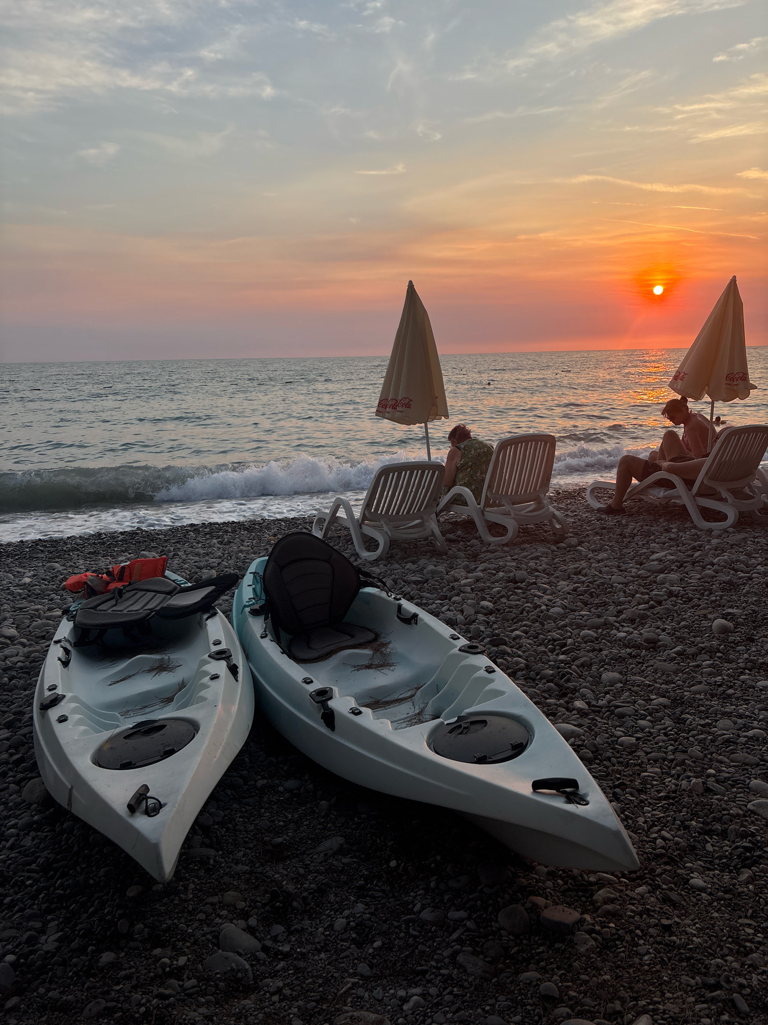A beach scene in Bar, Montenegro, at sunset. Two kayaks are in the foreground on a pebble beach. In the middle ground, several lounge chairs are set up with umbrellas. Two people are sitting on the lounge chairs, and the sun is setting over the Adriatic Sea. The sky is a soft pink and orange, with some clouds. The waves are gently lapping the shore. The overall mood is peaceful and serene.
