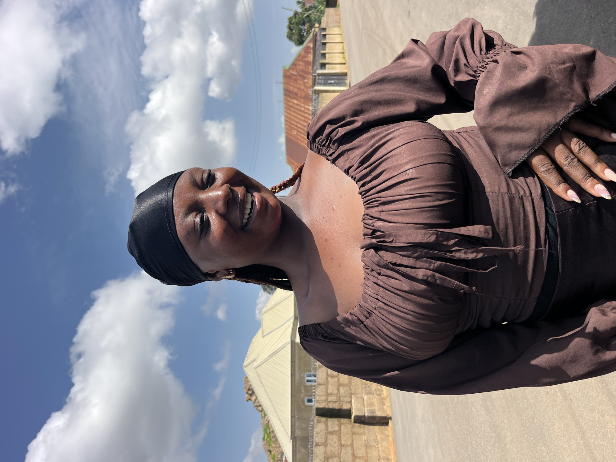 A young Black woman with dark skin, braids, and a black headscarf smiles down and to her right, her eyes closed. She is wearing a brown, ruched, long-sleeved top. The background shows a bright blue sky with fluffy white clouds, a stone wall, and buildings with corrugated roofs. She appears to be standing outdoors in a street or courtyard.
