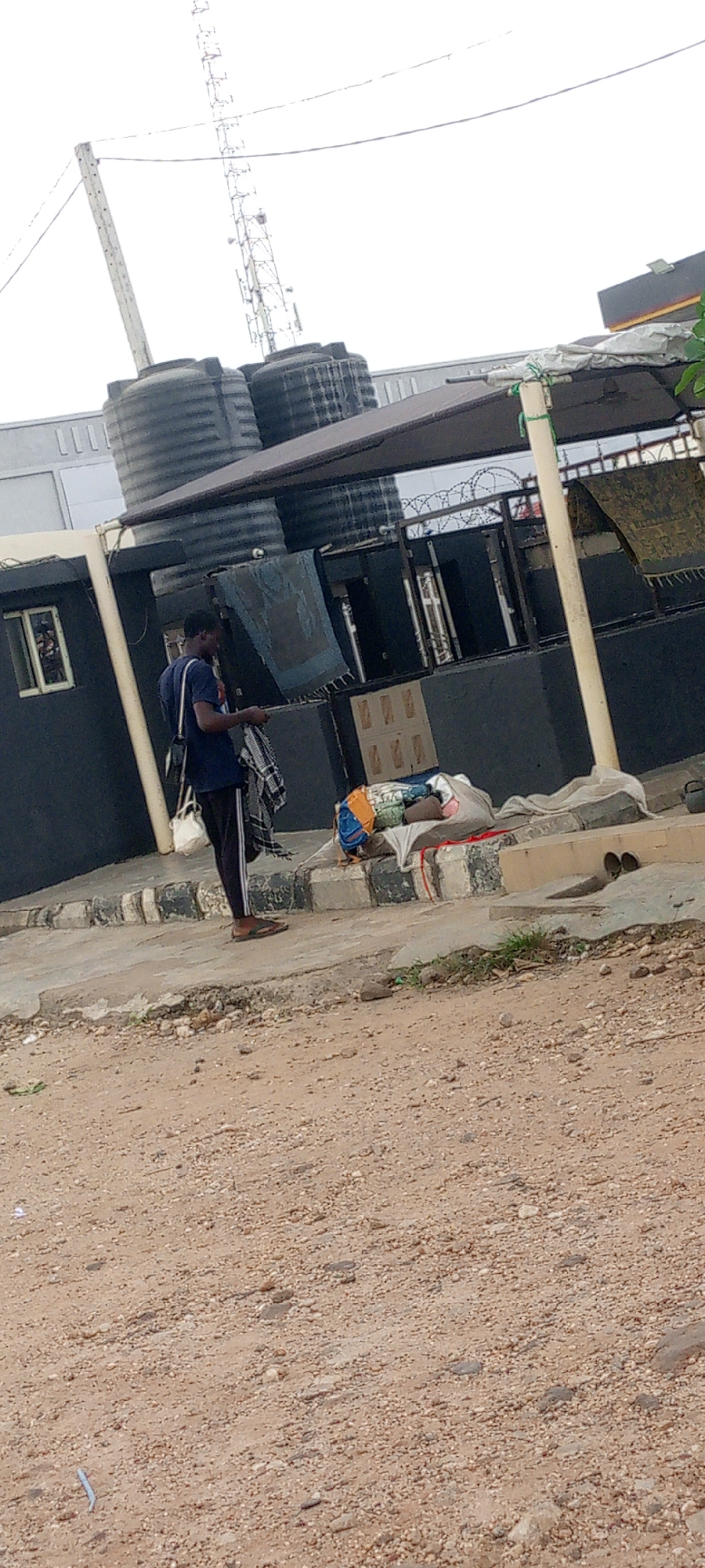 The image depicts a man standing near the side of a building with water tanks on the roof. He wears a dark blue t-shirt, dark pants with a light stripe on the side, and brown sandals. He carries a white bag on his shoulder and a patterned cloth in his hand. Next to him is a pile of bags and cloths on the ground. The building has a dark gray exterior and a small window. The area in front of the building appears to be unpaved with dirt and gravel. A cell tower is visible in the background. The location is suggested to be Zango-ndaji, Nigeria.