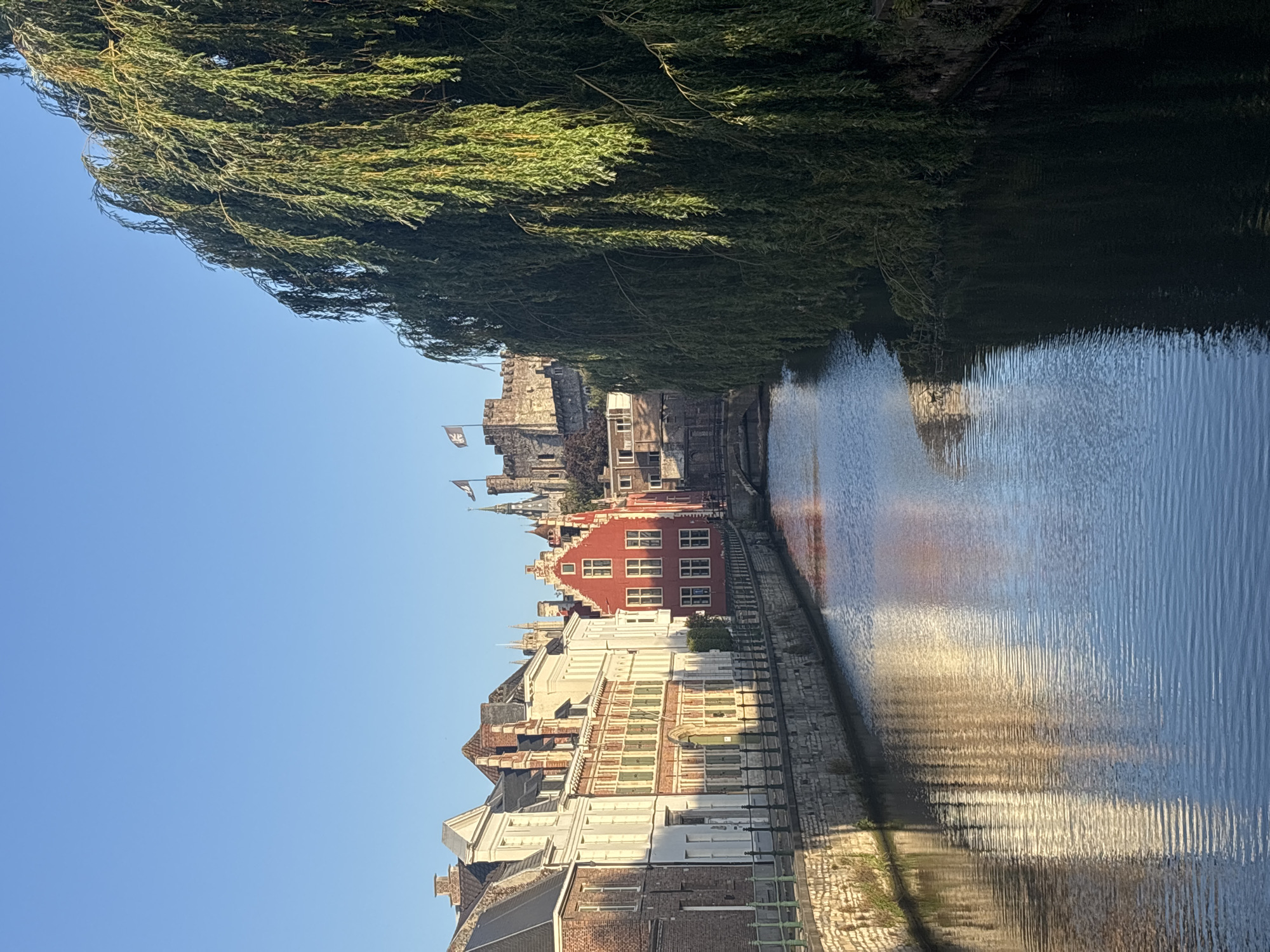 The image depicts a picturesque canal scene in the city of Ghent, Belgium, likely captured during the morning or late afternoon due to the warm, angled sunlight. The canal, with its calm, rippling water reflecting the surrounding architecture and foliage, forms the central element of the composition.
On the left bank of the canal, a row of traditional Flemish buildings lines the waterfront. These buildings exhibit varied architectural styles, including brickwork and plaster facades, with characteristic pitched roofs and multi-paned windows. Some buildings feature decorative gables and ornate stonework. A stone embankment with a railing separates the buildings from the canal.
Dominating the right side of the image is a large, mature weeping willow tree, its long, drooping branches creating a lush green curtain that partially obscures the view of the canal and the buildings beyond.
In the background, a medieval castle, possibly Gravensteen Castle, is visible perched on a hill, its stone battlements and towers silhouetted against a clear blue sky. Several flags, bearing dark emblems on a lighter background, fly from the castle. The overall impression is one of historical charm and natural beauty, characteristic of Ghent's historic center. No people are visible in the scene.