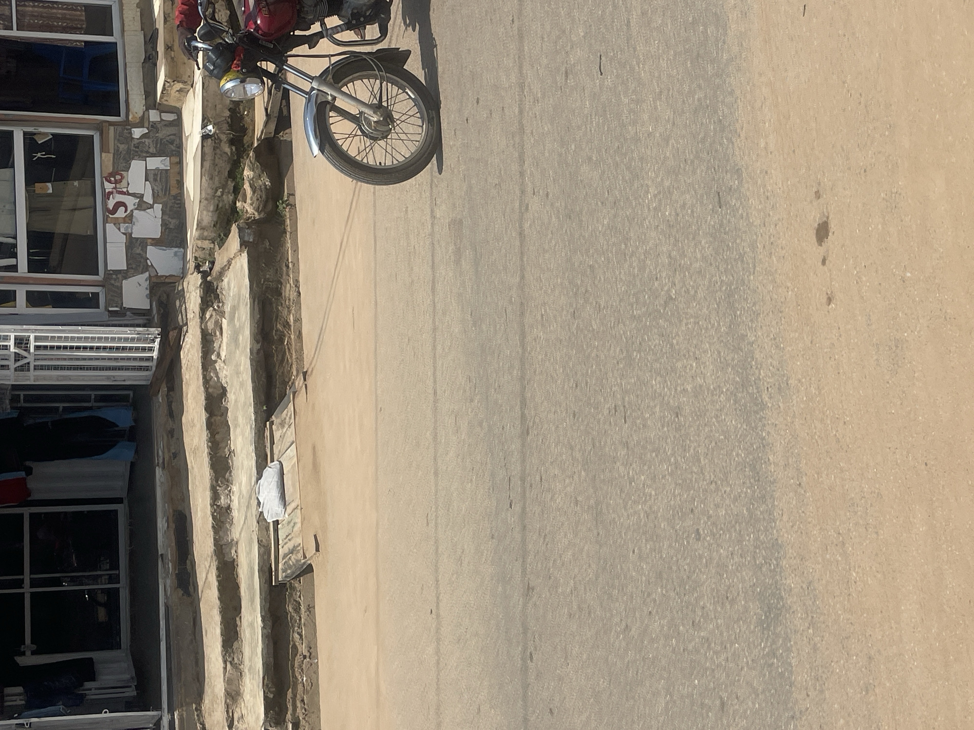 The image shows a street scene in Abuja, Nigeria. A red motorcycle is parked on the right side of the frame, facing left. The motorcycle's front wheel and part of its frame are visible. Behind the motorcycle, there are some steps leading up to a building. The building appears to be a shop, with its entrance open and displays visible inside. Clothing items are hanging in the shop entrance. The steps are made of concrete and show signs of wear and tear. The street itself is paved with asphalt and has a dusty appearance. The overall environment suggests an urban setting, likely in a commercial area.