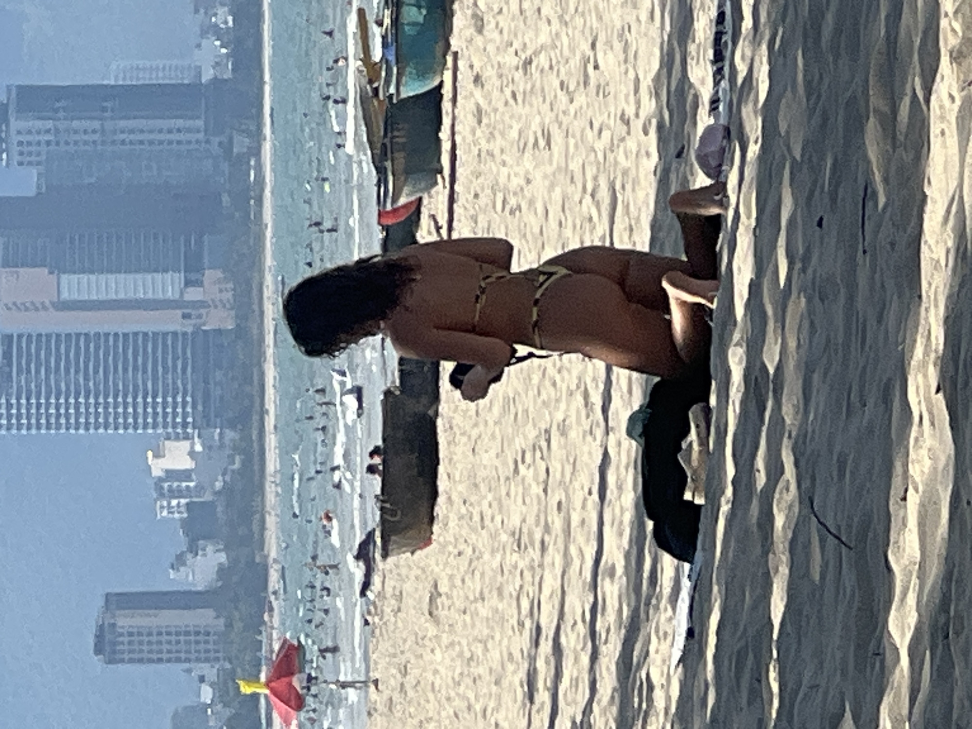 A person is kneeling on a sandy beach, facing away from the camera, wearing a bikini. They appear to be holding a camera or phone. In the background, the ocean is filled with swimmers and small boats. Further in the background, a cityscape with tall buildings and a hazy sky is visible. The scene is likely on a beach in Da Nang, Vietnam, given the architecture of the buildings and the presence of traditional Vietnamese fishing boats. The lighting suggests it is daytime.