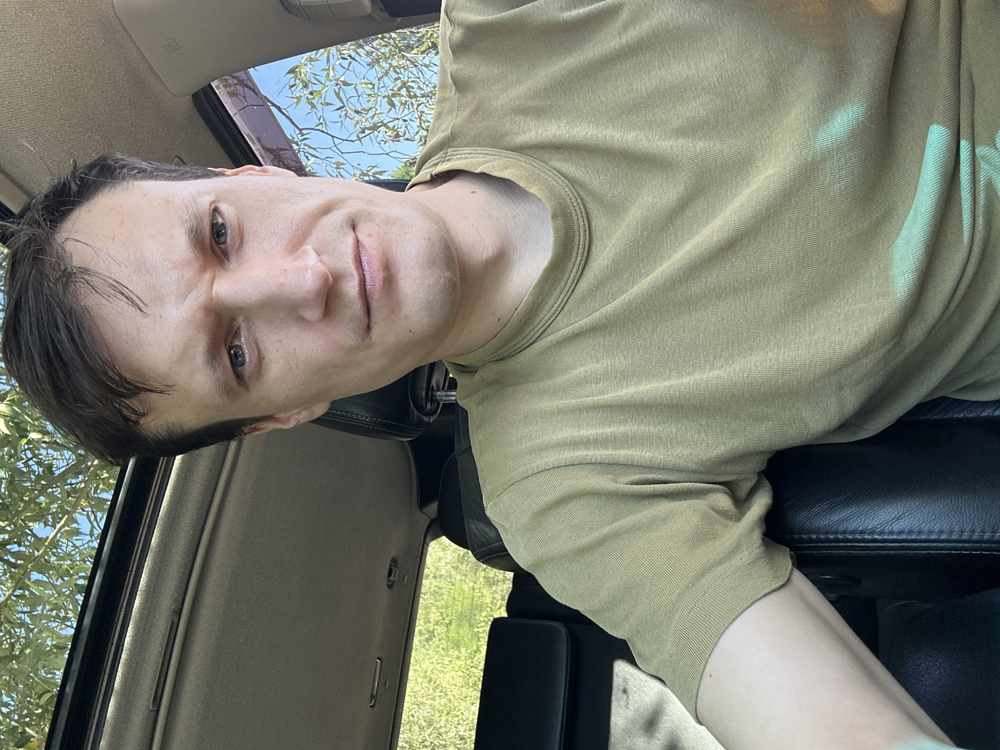 A man is sitting in the driver's seat of a car, looking at the camera. He has brown hair and is wearing a green t-shirt. The car has a sunroof, which is open, and trees and a blue sky can be seen through it. The interior of the car is dark, with black leather seats visible. The man's left arm is resting on the armrest. The image was taken outdoors in Makiv, Ukraine, as suggested by the presence of trees and the type of vehicle common in the area.