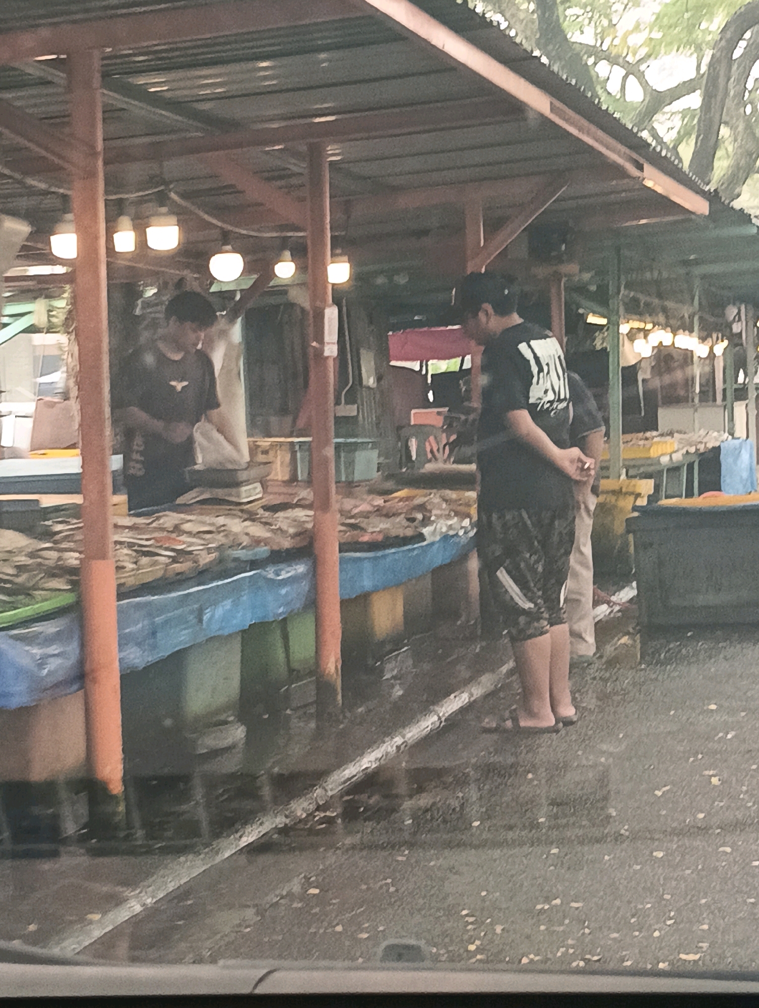 The image depicts a market stall, likely an outdoor fish market, in Kuching, Malaysia. Two individuals are visible. One person, wearing a dark t-shirt and patterned shorts, stands facing away from the camera, with hands clasped behind their back. The other individual, wearing a dark t-shirt, is partially obscured and appears to be interacting with a scale on the stall. The stall is laden with dried fish arranged on a blue tarp. Wooden posts support a corrugated metal roof overhead, with lighting fixtures present. The ground appears wet, suggesting recent rain or a damp environment common in tropical markets. The background shows other market stalls and trees.