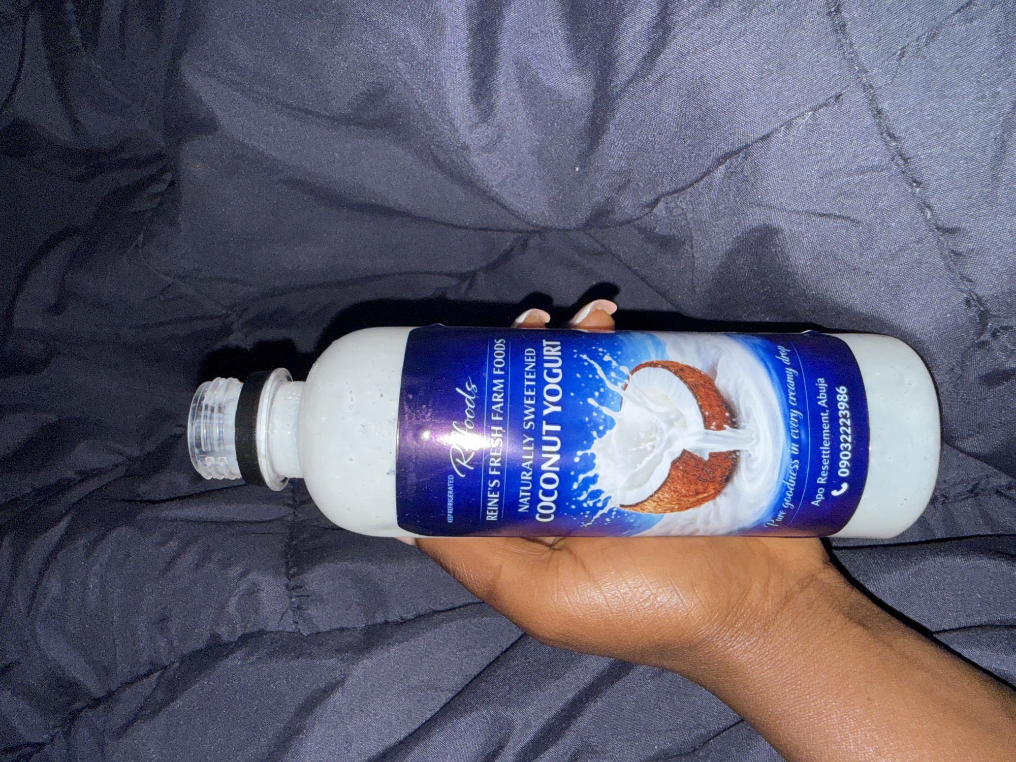 A person's hand is holding a plastic bottle of Reine's Fresh Farm Foods Naturally Sweetened Coconut Yogurt. The bottle has a blue label with white text and a picture of a coconut with milk being poured over it. The text on the label indicates the product is from Apo Resettlement, Abuja, and provides a phone number: 09032223986. The background is a dark, textured fabric, likely bedding, and appears to be indoors.