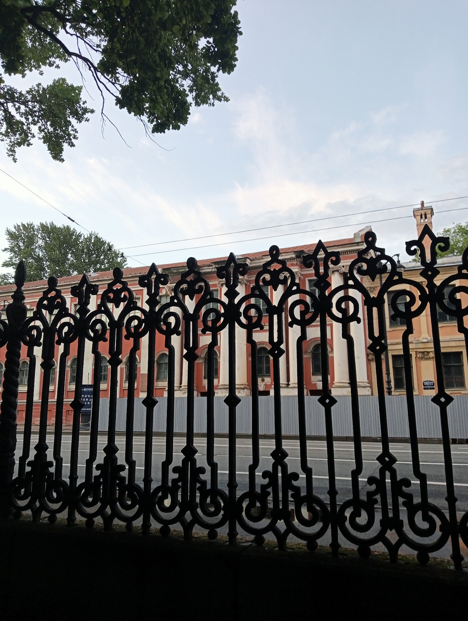 The photograph captures an ornate black metal fence in the foreground, silhouetted against a pale sky with wispy clouds. Behind the fence, a historic building with red and white facades is visible, featuring arched windows and classical architectural elements like columns and decorative cornices. A section of a street with visible white lines runs horizontally in the middle ground, partially obscured by the fence. Trees with green foliage are present in the upper left and upper right of the frame. Electrical wires are strung across the sky. The scene suggests a view of an architectural landmark in Saint Petersburg, Russia, likely a public institution or a historical building.