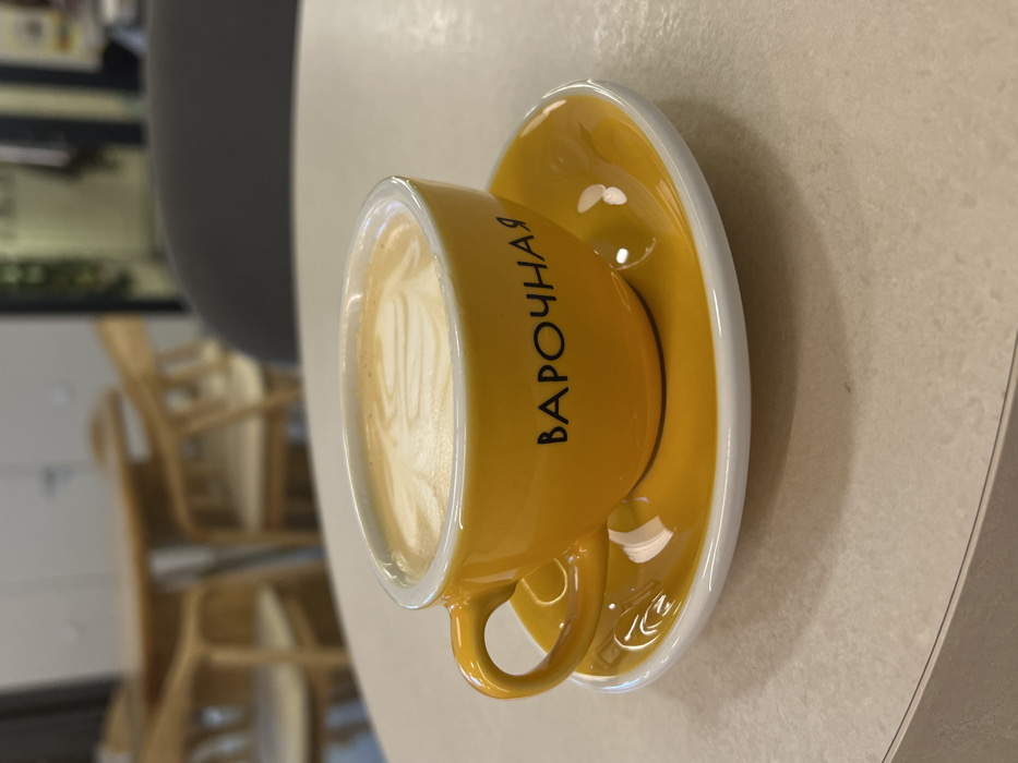 A bright yellow ceramic coffee cup with a matching saucer is centrally positioned on a light-colored, speckled table surface. The cup contains a beverage topped with white frothed milk, exhibiting latte art. Black Cyrillic text, "ВАРОЧНАЯ", is visible on the side of the cup. The background is blurred, showing parts of multiple light-colored wooden tables and chairs, alongside a dark grey chair back, indicating an indoor cafe or coffee shop setting. No individuals are visible. Based on the branding "ВАРОЧНАЯ" (Varochanaia), the location is a Varochanaia coffee shop in the city of Krasnoyarsk, Russia.
