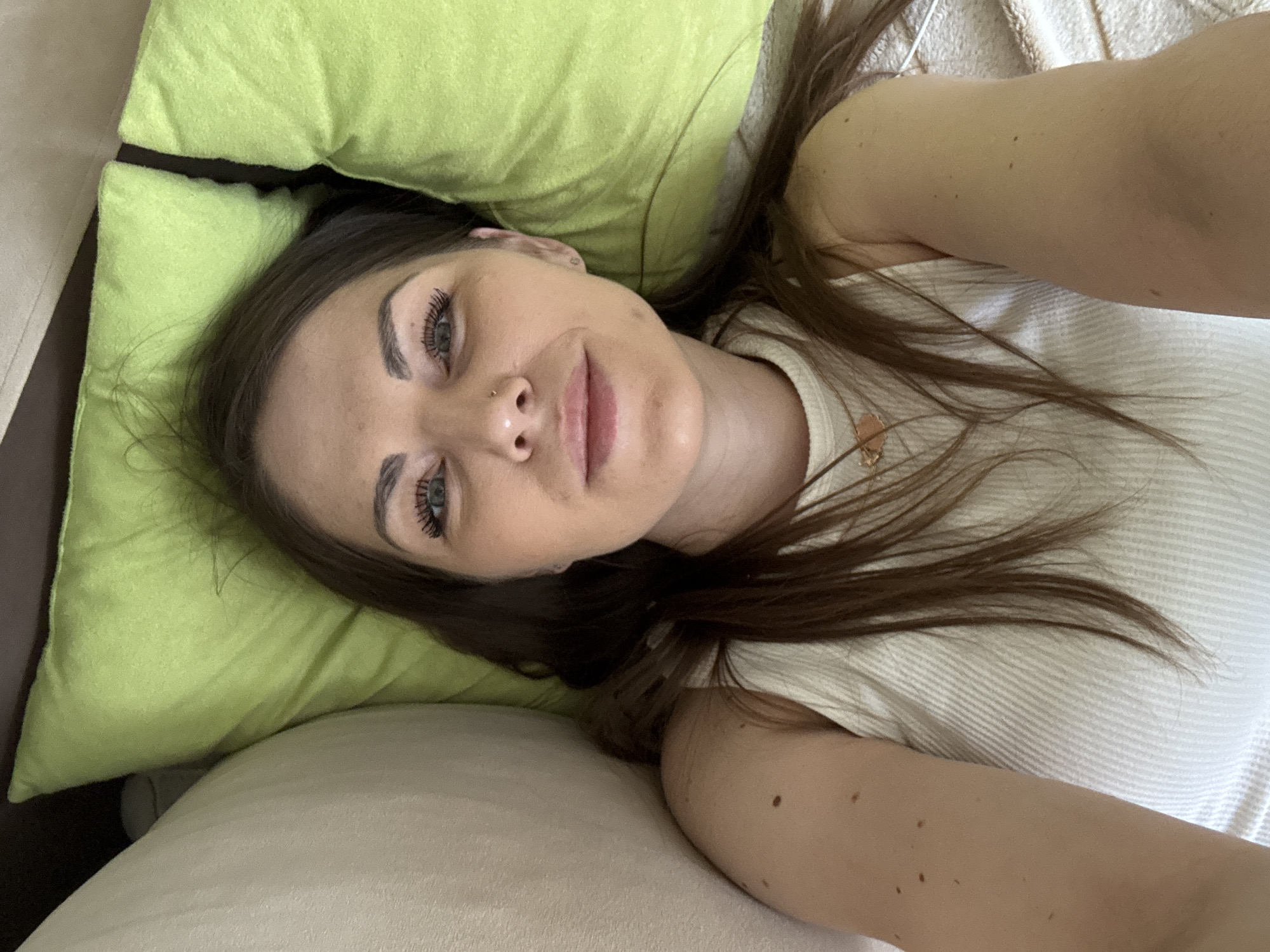 A woman lies on a bed. Her head rests on a green pillow. The woman has brown hair and a nose ring. She is wearing a white shirt. The bed has white sheets. There is no evidence that the image was taken in Razdolye, Russia.