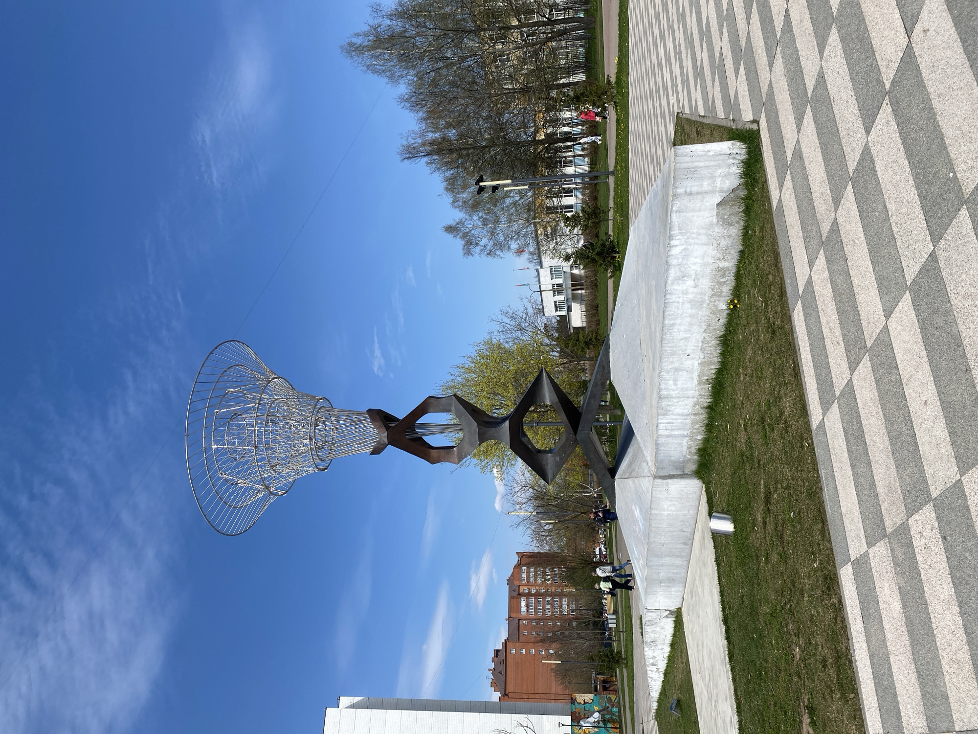 The image shows a vertical view of a public space in Unknown, Unknown. A tall, modern sculpture dominates the foreground, consisting of a wire mesh cone shape atop a series of dark metal geometric shapes connected to a concrete base. Pedestrians walk along a pathway beside the sculpture. Behind the sculpture, several multi-story brick and white buildings are visible. Bare trees stand behind the buildings. The sky is a clear blue with scattered white clouds. The ground near the sculpture is covered in grass, and the walkway is paved with alternating gray and white squares.