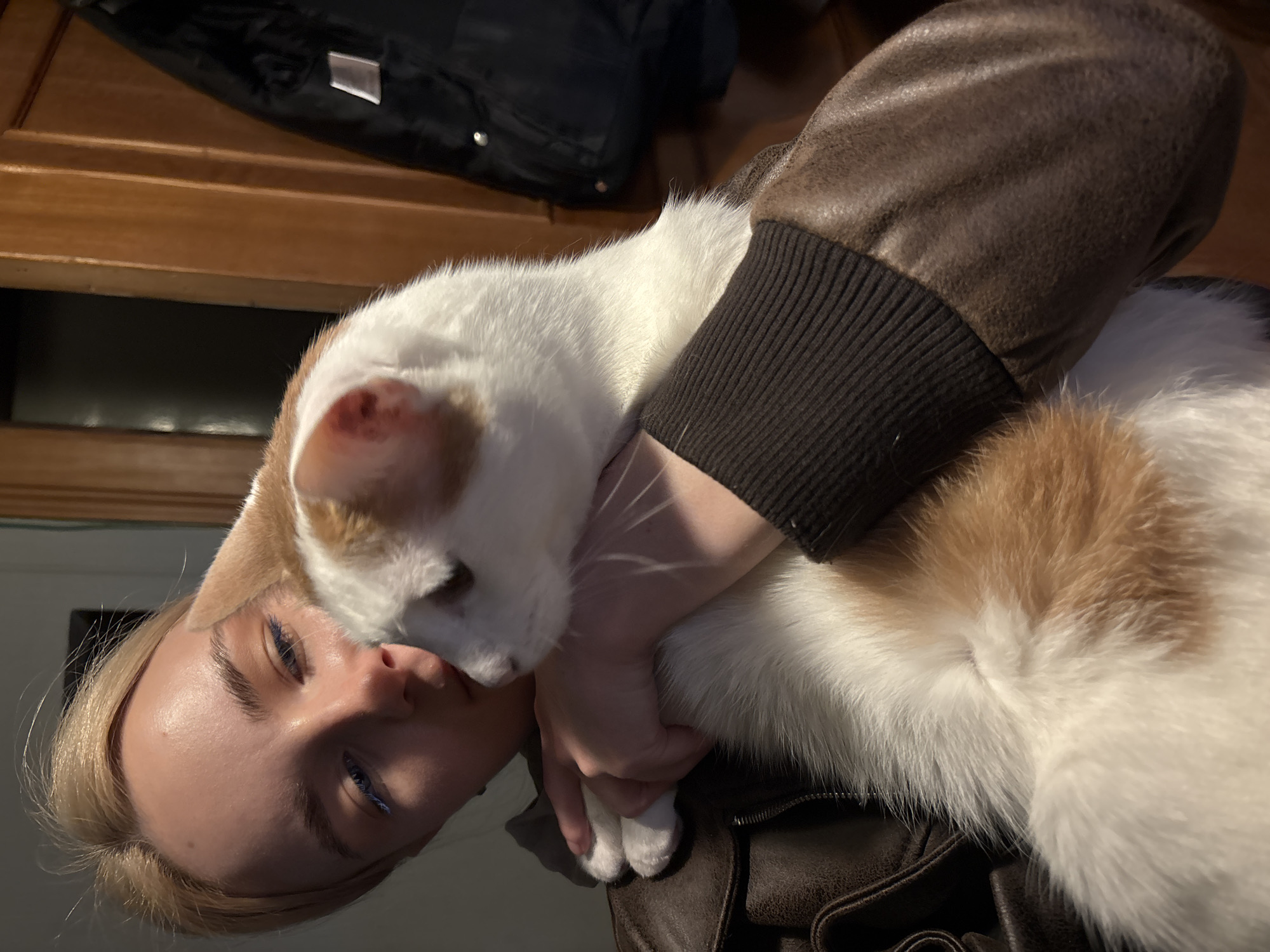 A young Caucasian person with blonde hair, wearing a brown leather jacket, is holding a white and orange cat. The person's face is partially obscured by the cat as they look down at it. The cat is nestled against the person's chest and shoulder, its head turned to the side. The background appears to be an indoor setting with wooden paneling and a dark jacket hanging on the wall. The lighting suggests an indoor environment, possibly in a home. The location is Novoe Devyatkino, Russia.