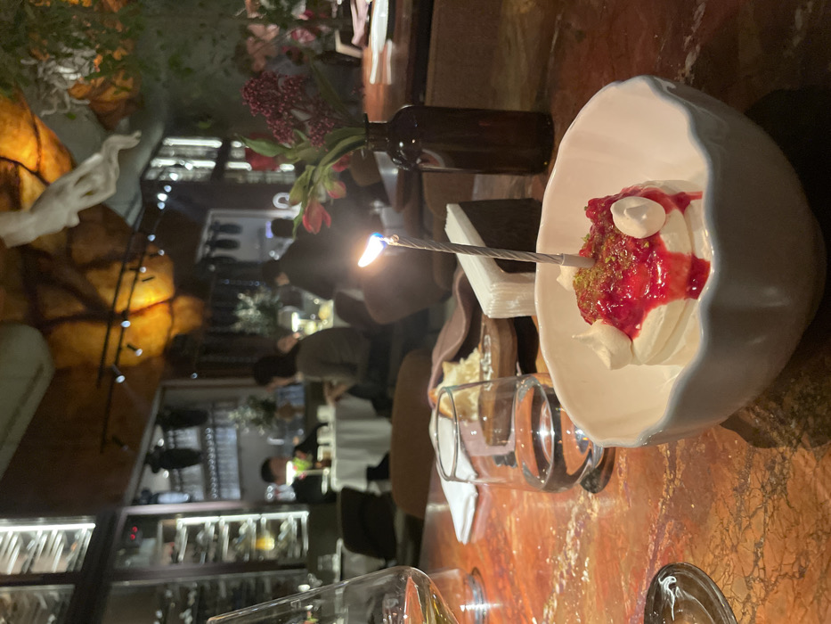 A dessert, featuring white cream, red syrup, and green sprinkles, is presented in a white, shell-shaped bowl with a lit birthday candle. The dessert is placed on a wooden table next to a glass. In the background, blurred figures and warm lighting indicate a restaurant setting with decorations. A vase with red flowers and a dark bottle add to the table setting.