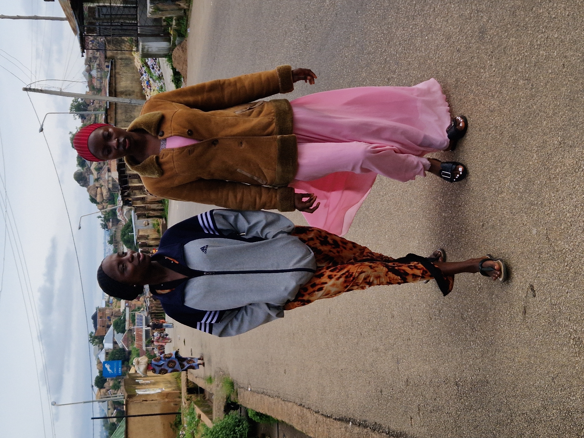 Two young women walk on a paved road in Kabong, Nigeria. The woman on the left wears a grey Adidas jacket with blue trim and black and white stripes on the sleeves, over a leopard-print skirt and black sandals. She has a black head covering. The woman on the right wears a brown fuzzy jacket with a fur collar over a pink dress and black sandals. She has a red head covering. In the background, there are buildings, a street lamp, and a hillside with more buildings. The sky is cloudy.