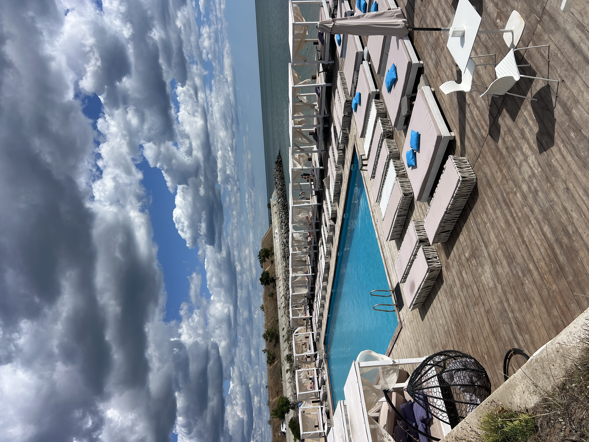 This is a wide-angle, eye-level, outdoor, full-shot photograph of a resort area in Fontanka, Ukraine, on a partly cloudy day. The scene depicts a luxurious beach club with a swimming pool and numerous sun loungers. The swimming pool is rectangular and filled with bright blue water. It is surrounded by a wooden deck. Along the sides of the pool and extending further back are rows of white cabanas, each with white curtains.
In front of the cabanas are rows of beige sun loungers, some of which have bright blue cushions. There are a few small white tables and chairs set up on the deck. In the foreground on the left, there is a black hanging egg chair with gray cushions.
Beyond the resort area, there is a body of calm, dark blue water, likely the Black Sea, stretching to the horizon. A rocky embankment and a sloped grassy hill with sparse vegetation can be seen in the background. The sky is filled with a mix of dark gray and white clouds against a clear blue sky. There are a few people visible in the distance near the water and within some of the cabanas, but they are not the primary focus of the image. The overall impression is one of a relaxed and upscale vacation destination.