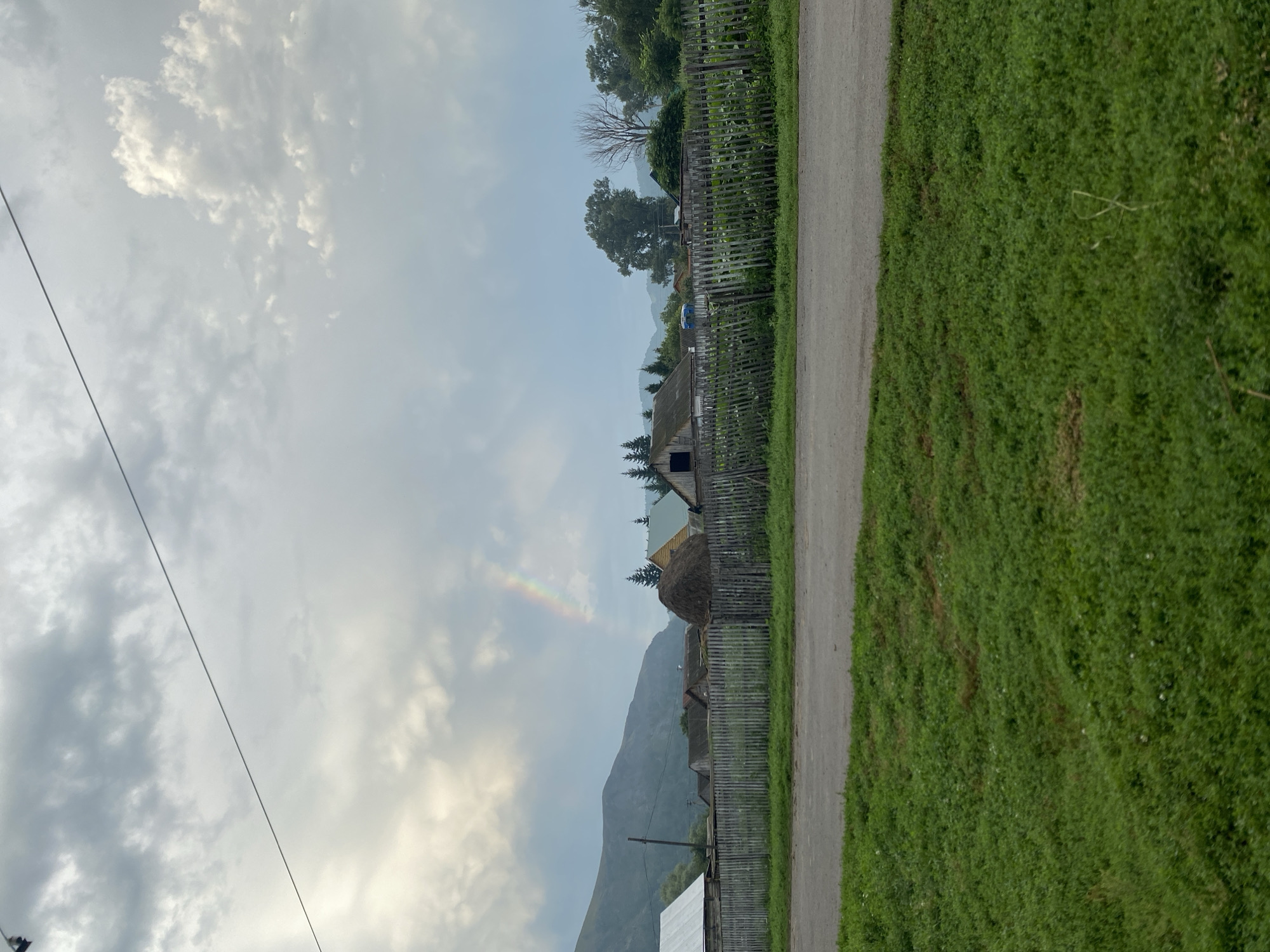 The image captures a rural scene in Maloarslangulovo, Russia, under a cloudy sky with a faint rainbow visible. In the foreground, a paved road separates a strip of green grass from a wooden fence. Beyond the fence lies a collection of village houses, some with visible roofs and structures. A large haystack stands near the center, and trees and mountains form the backdrop. The overall atmosphere is subdued, with the cloudy weather and muted colors contributing to a sense of calm. There are no people or animals present in the frame.
