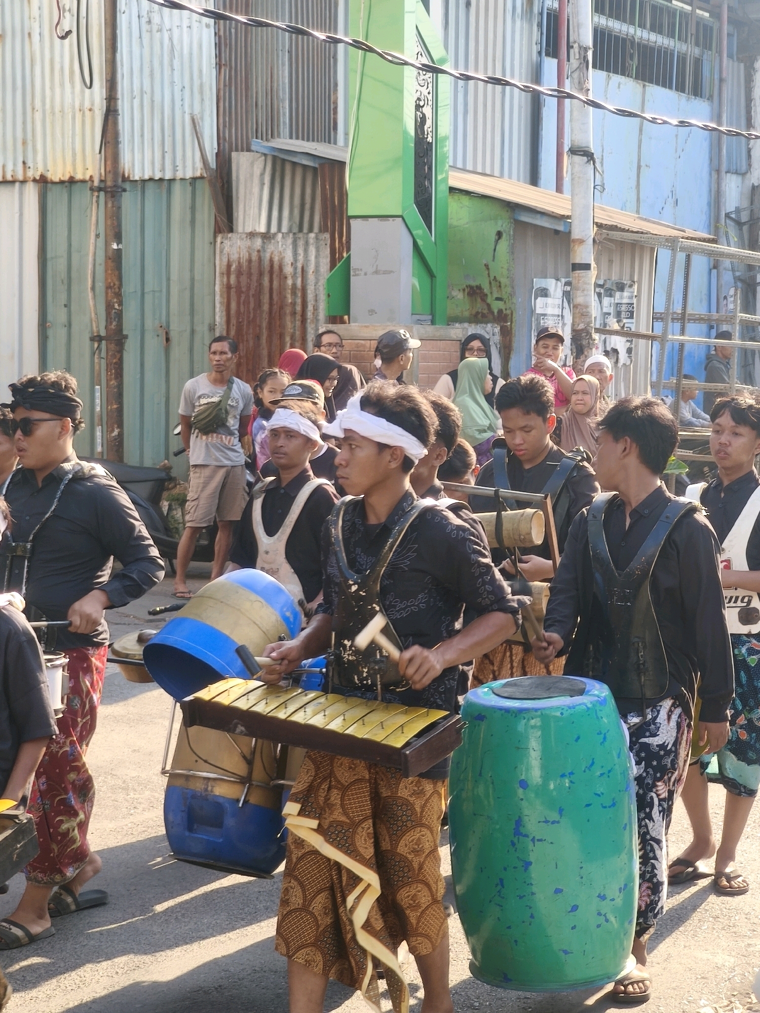 This is a vertical, full-shot photograph taken at eye-level of a street procession in Kepuh Kiriman, Indonesia. The scene features a group of young men, some of whom are wearing traditional Indonesian clothing and headbands, playing percussion instruments. The man in the foreground on the left, wearing a black shirt and sunglasses, is holding a drumstick and appears to be playing a metal xylophone. To his right, another young man in a black shirt is playing a gamelan. Behind them, a large blue and yellow drum is visible. Further to the right, a young man in a black shirt and white headband is playing a large green drum. The procession is taking place on a paved street. The background consists of various buildings and structures made of corrugated metal and painted surfaces, suggesting a somewhat impoverished urban or semi-urban environment. The people in the background are a mix of men, women, and children, some of whom are watching the procession. The lighting indicates it is daytime, likely late afternoon given the long shadows cast by the people. The overall atmosphere suggests a cultural event or celebration.