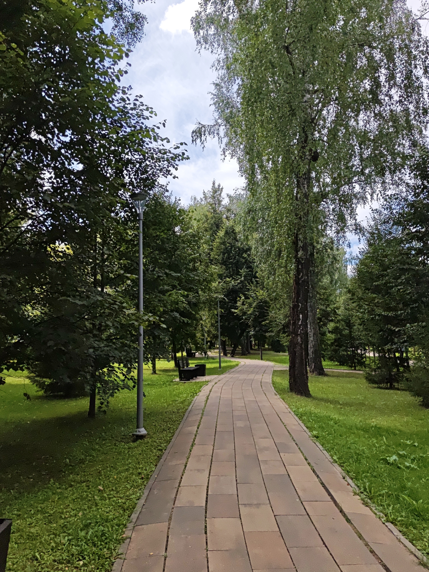 The image depicts a paved pathway winding through a park with lush green grass and numerous trees. The pathway, made of rectangular stone tiles, curves to the right, disappearing into the distance. On either side of the path, there are benches, suggesting a public recreational area. Tall trees, including birches with distinctive white bark, dominate the landscape, casting shadows on the grass. The sky above is partly cloudy, with white clouds scattered across a pale blue expanse. There are no people visible in the frame. The scene suggests a peaceful, well-maintained park environment, likely in a suburban or urban setting. The location is identified as Shcherbinka, Russia, a town in the Troitsky Administrative Okrug of Moscow.