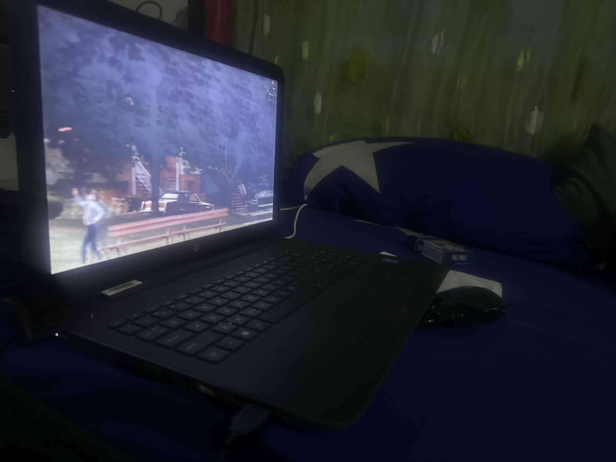 An indoor scene captured in Jos, Nigeria, features a black HP laptop positioned on a dark blue surface, likely a bed or mattress. The laptop screen is illuminated, displaying a blurry image of what appears to be a video game or a film. The digital scene shows a person with their arm raised, a reddish-brown structure, a dark vehicle, and trees under a dimly lit sky.
To the right of the laptop, a dark blue pillow adorned with a large white star rests against a green background, possibly a curtain or fabric. A pack of "PALL MALL" cigarettes is visible next to the pillow. A black computer mouse is partially obscured near the cigarette pack. The lighting in the room is dim, suggesting it is either evening or night, with the laptop screen serving as a primary light source. The overall impression is of someone relaxing or unwinding, possibly playing games or watching media in a personal space.
