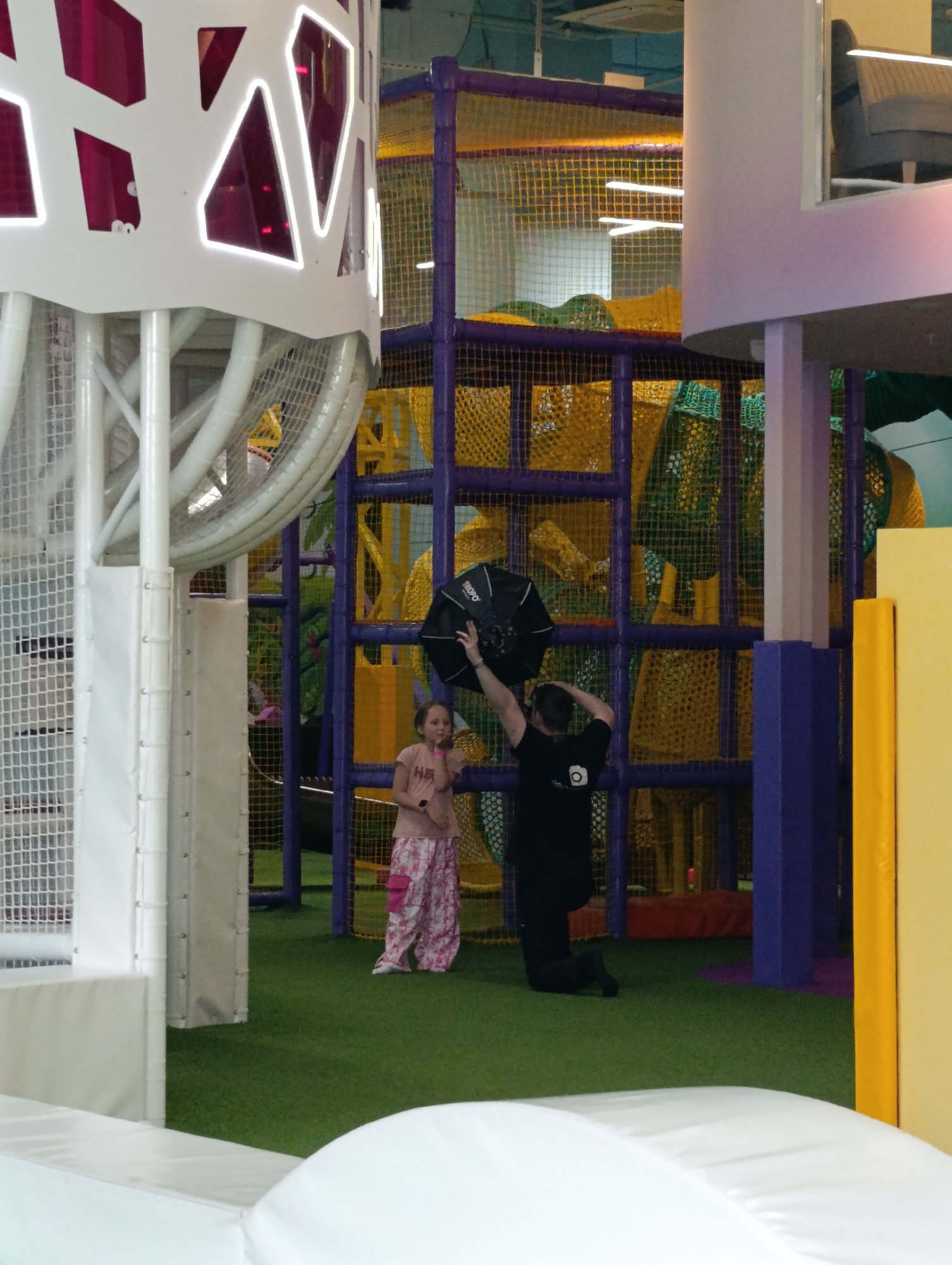 In Moscow, Russia, an indoor playground area features a young girl in a pink t-shirt and patterned pants standing on green turf. To her right, a man in a black t-shirt and dark pants is kneeling, holding a black umbrella-like photography light modifier above his head. The background consists of a multi-level play structure with yellow and green netting and purple support poles. A white structure with illuminated pink geometric shapes is visible on the left.