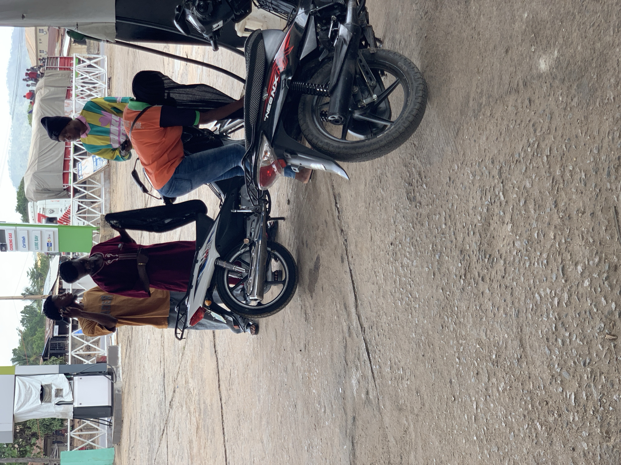 This image captures a daytime scene at a gas station or roadside service area in Lokoja, Nigeria, with several individuals and motorcycles present. The ground is a mix of concrete and gravel.
In the foreground, two motorcycles are parked. On the left, a man wearing a brightly colored, multi-patterned long-sleeved shirt (yellow, green, blue, pink), a dark beanie, and blue jeans stands next to a black motorcycle. Another man, wearing an orange short-sleeved shirt with "PRIDE" partially visible on the back and blue jeans, is seated on this black motorcycle, which features "NEBNX" in red lettering on its side. To the right, a second motorcycle, primarily white and grey, is parked. Standing near this motorcycle is a man in a maroon long-sleeved top with white embroidery and dark trousers, while another man behind him, in a yellow-brown t-shirt, is engaged in a phone call.
The background shows typical gas station infrastructure, including white and green gas pumps. Visible signs advertise services such as "COOKING GAS," "Car Wash," and "LUBE BAY," alongside "24/7." A large white truck with a covered bed is parked further back, and a group of people can be seen gathered around it. Distant hills or mountains are faintly visible under a clear sky. The scene depicts a casual stop, possibly for refueling or a break.