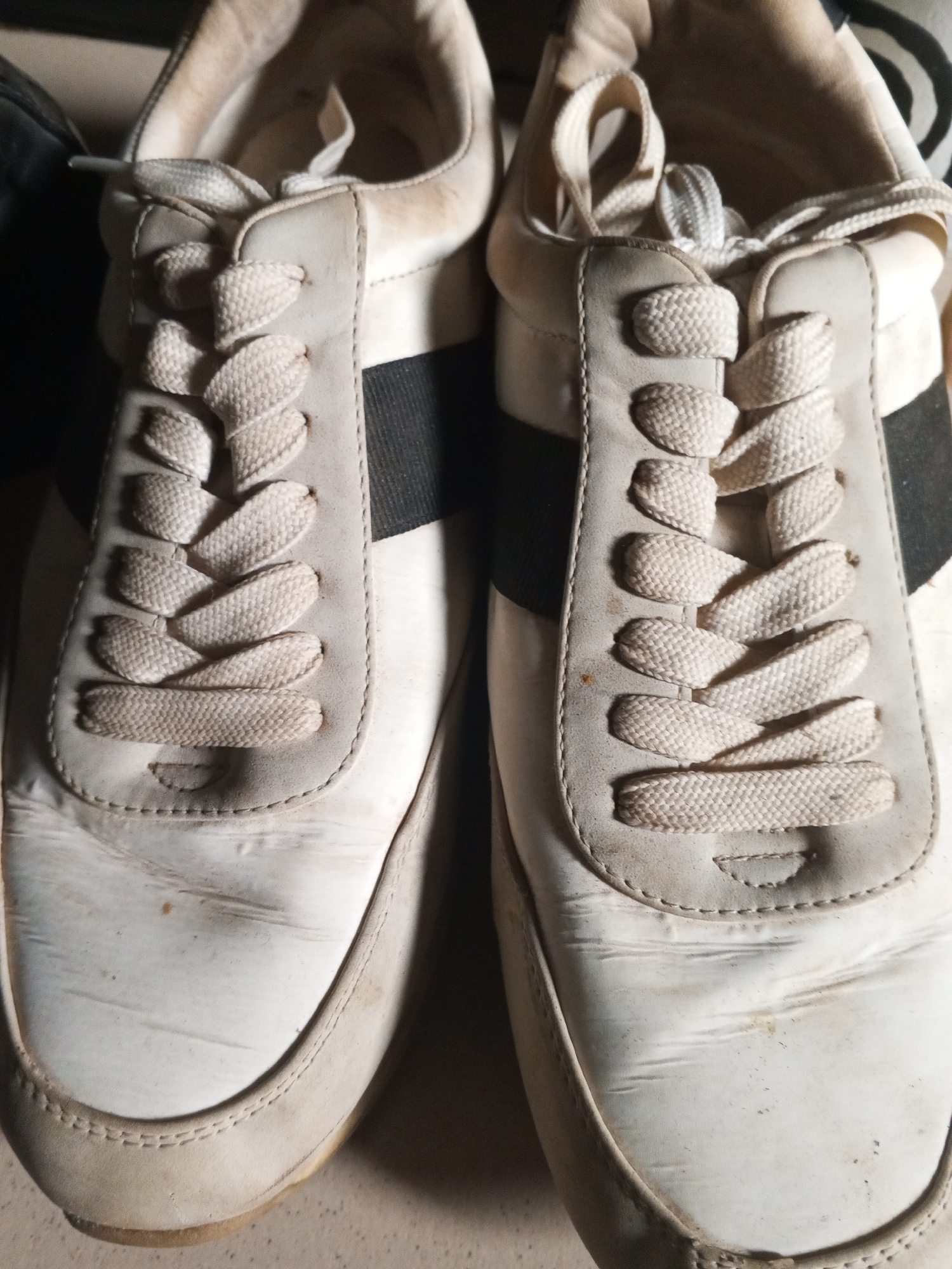 The image shows a close-up, overhead view of a pair of white sneakers. The sneakers appear to be made of leather and have a worn, slightly dirty appearance with visible creases and scuff marks. Each shoe has white laces threaded through eyelets. There is a black stripe across the side of each shoe, just above the sole. The background is dark and out of focus, providing no discernible context for the scene. The lighting is dim, suggesting an indoor setting or low light conditions. There is no text visible in the image. The focus is on the shoes themselves, highlighting their condition and design details. The overall impression is of a pair of well-used casual shoes.