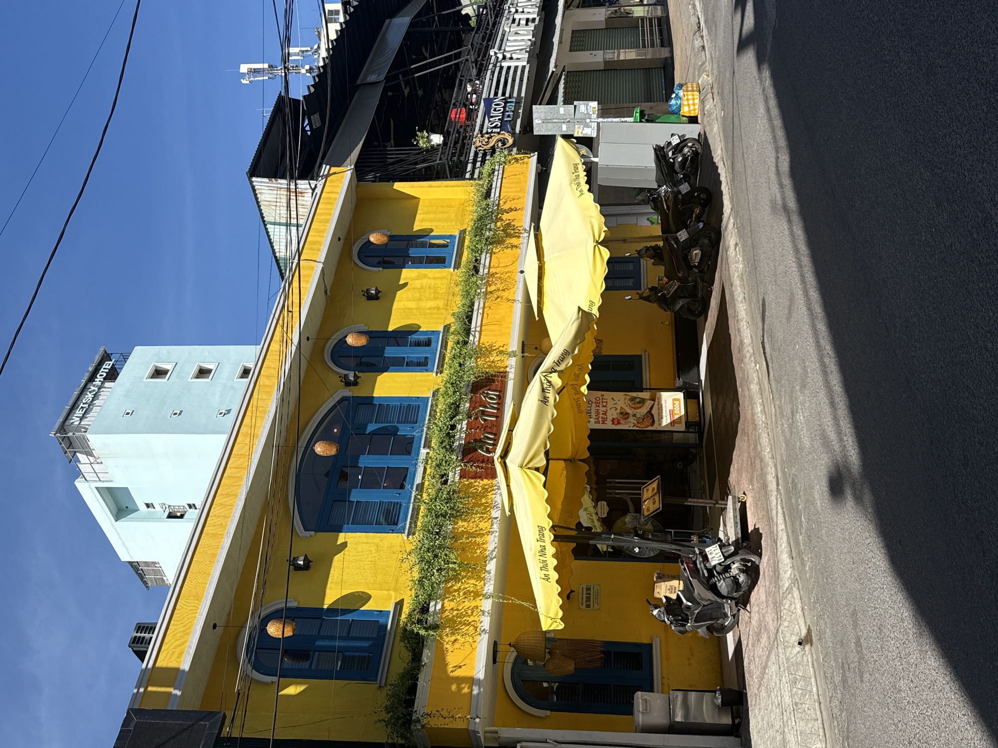 The image shows a vibrant yellow building with bright blue window frames. The building appears to be a restaurant called "Ăn Thôi Nha Trang", as evidenced by the signs and awnings displayed. The awning is bright yellow and provides shade to an outdoor seating area. Hanging plant vines decorate the exterior of the building between windows. Street-parked motorbikes are visible in front of the restaurant along the edge of the street. In the background, a light blue building with signage visible that says "Viet Sky Hotel". Across the street are other signs that say Saigon Grill. The sky is a clear blue. The location is in Nha Trang Ward, Vietnam.