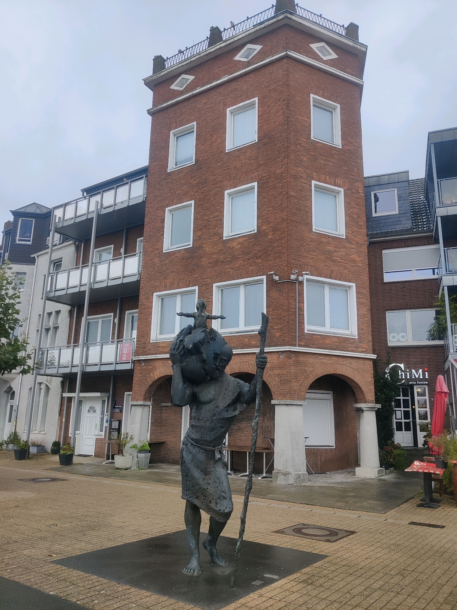 The image shows a bronze statue of a male figure carrying a globe on its head, with a stick in its right hand, positioned in a paved square in front of brick buildings in Emmerich on the Rhine, Germany. The statue stands on a dark rectangular base. The buildings feature multiple stories with balconies and windows. To the right of the statue, an arched entrance leads to a business labeled "ChiMi asia fusion" with red umbrellas and tables outside. The sky is overcast, and the ground is wet, suggesting recent rain. The overall scene depicts an urban setting with public art and commercial establishments.