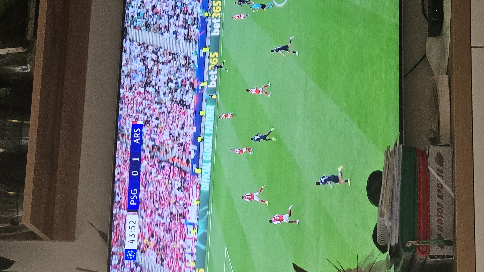 The image depicts a television screen displaying a soccer game between PSG and ARS (Arsenal). The score is 0-1 in favor of Arsenal at the 43:52 minute mark. The game is being shown with "bet365" as an "OFFICIAL GLOBAL PARTNER".
The setting appears to be indoors, as the television sits on a surface that includes stacked papers and a case labelled "MOTOR SPO/8". The exact location within Plovdiv, Bulgaria, is not discernible from the image. There is no information available to determine a location in Plovdiv, Bulgaria.