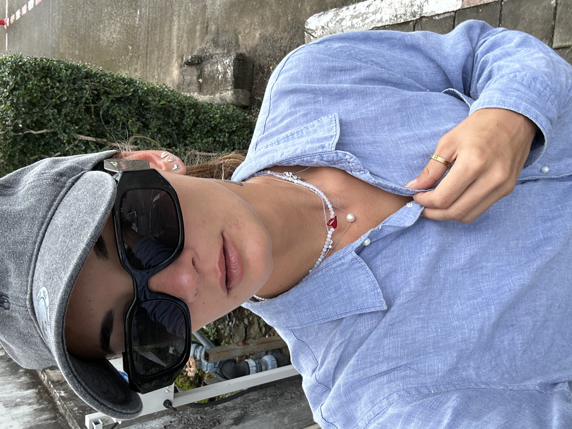 A young woman wearing a gray baseball cap, black sunglasses, and a light blue button-down shirt stands outdoors. She has on a delicate necklace with pearls and a red heart charm, and a gold ring on her right hand. Her left hand is buttoning her shirt. The background shows a concrete path, a dense green hedge, and some outdoor plumbing or pipes. The scene is likely on a street or walkway in Kathu, Thailand, given the tropical vegetation and outdoor setting.