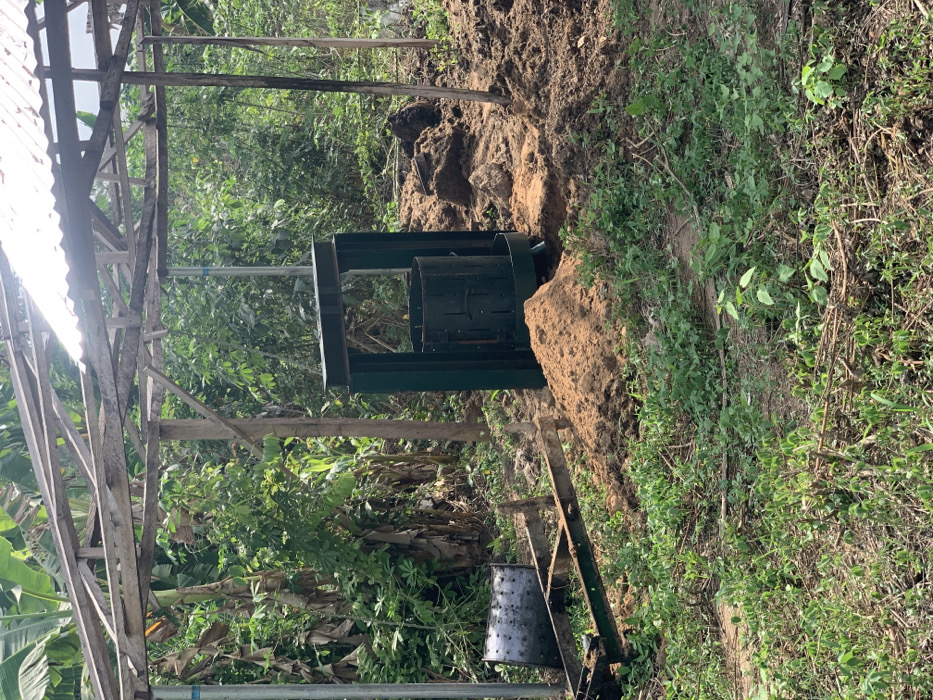 A dark green compost bin with a metal frame and a metal drum is situated in a small, unpaved area in a backyard in Egbeda, Nigeria (latitude: 7.12, longitude: 5.362). There is a pile of dirt next to the bin. Behind the bin is a tall, leafy, green tree. There is a wooden structure with a corrugated metal roof. There are green plants growing in the area. The ground is covered in dirt and leaves. The bin is tilted slightly to the right. The metal drum is partially open, and a small amount of dirt is visible inside. The area appears to be relatively unused, with signs of minimal activity.