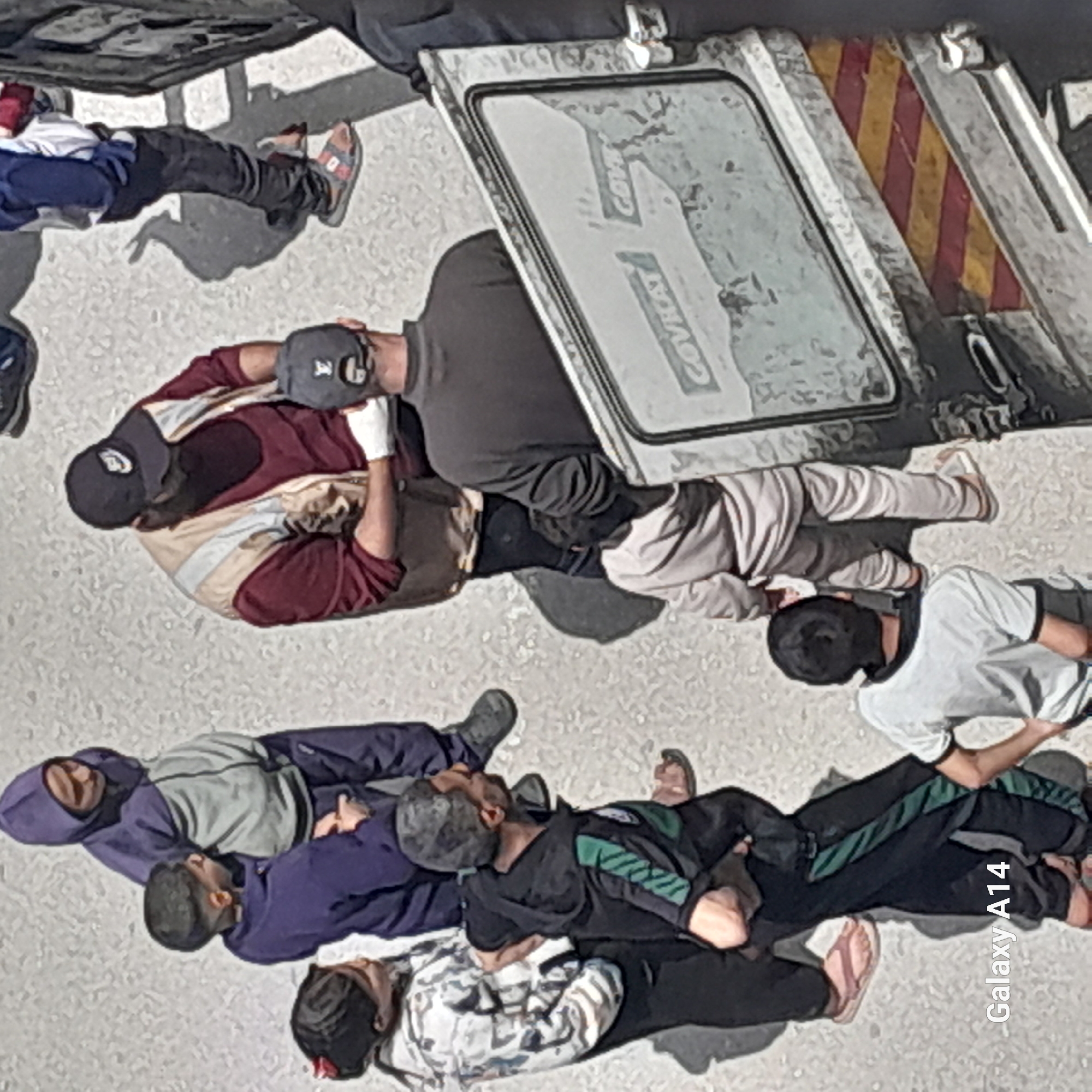 The image presents an overhead view of at least nine individuals lying on a light-colored, possibly paved or concrete surface. Most subjects appear to be adult males, dressed in casual wear including t-shirts, hoodies, pants, and caps. Some wear sandals, while others have sneakers. Their positions vary, with some lying face down, others on their backs or sides, suggesting a state of rest, exhaustion, or incapacitation.
In the upper portion of the frame, part of a large vehicle, possibly a truck or trailer, is visible. It features a metallic surface with a window-like panel on which faint, fragmented text, possibly "COVRCA," can be discerned. Adjacent to this panel, a distinct red and yellow diagonal striped pattern, characteristic of a hazard or warning marking, is visible on the vehicle.
The bright lighting and long, defined shadows cast by the subjects and the vehicle indicate that the photograph was taken during daytime, likely under sunny conditions. The environment appears to be an outdoor setting, possibly an urban or industrial area. The overall scene conveys a sense of stillness and inactivity among the group of people. In the bottom right corner of the image, the text "Galaxy A14" is visible, indicating the device used to capture the photo.