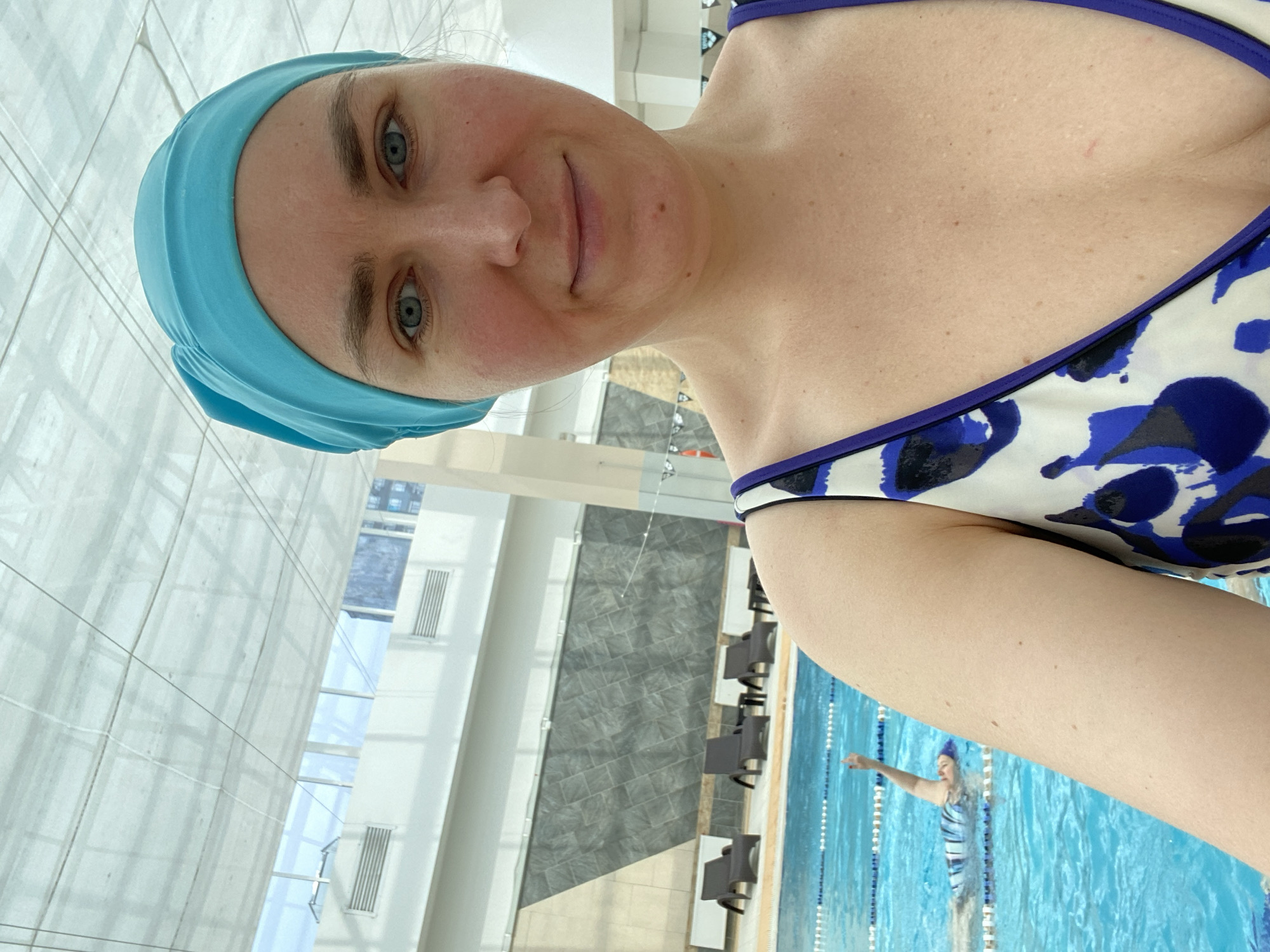 An indoor swimming pool facility in Yekaterinburg, Russia, features a woman taking a selfie. She is wearing a blue swimming cap and a swimsuit with a blue and white abstract pattern. The pool water is blue, and lane lines are visible. In the background, other swimmers are present in the water. The pool area is surrounded by lounge chairs and a wall with a decorative stone-like pattern. Above, a translucent ceiling structure allows natural light to filter in.