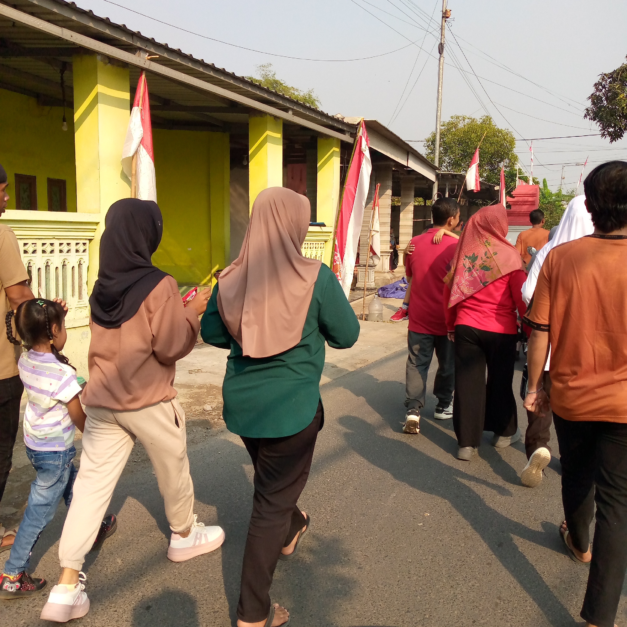 In Tambakagung, Indonesia, a group of people walk on a paved road in front of a yellow building with a metal roof. Several Indonesian flags, red and white, are displayed on poles along the building's facade and in the distance. The group consists of individuals of various ages, including women wearing headscarves and one child with braided hair. They are dressed in casual attire, with some wearing t-shirts and pants, and others wearing hijabs and loose-fitting clothing. The sun casts long shadows on the road, indicating it is either morning or late afternoon. The environment appears to be a residential area, with power lines visible overhead.