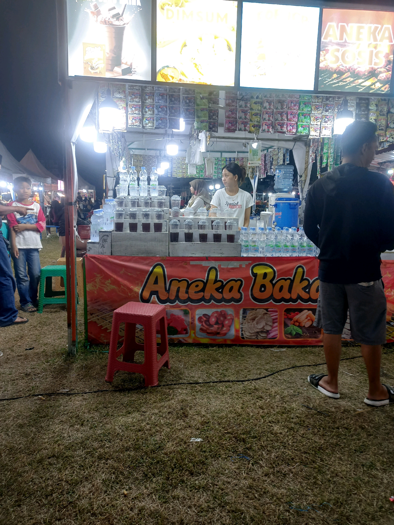 The image shows a street food stall at night in Tanon, Indonesia. The stall is selling drinks and snacks, with a banner displaying "Aneka Bakar" which translates to "Assorted Grills". Several people are present, including a vendor behind the counter, a customer in the foreground, and a young boy to the left. The stall is decorated with various products and illuminated by bright lights. The ground is covered with dry grass, suggesting an outdoor setting.