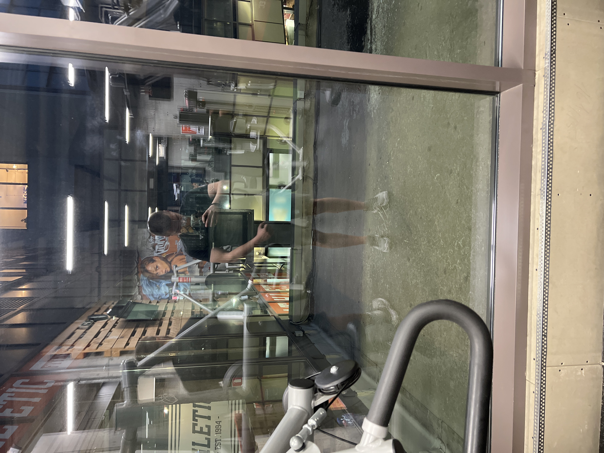 The image is taken from inside a building, looking through a window at a street or outside area. The reflection in the window obscures the view. The reflection includes a person holding a phone or camera. A piece of gym equipment is in the foreground. Visible through the reflection is the word "ATHLETIC." While "ATHLETIC" is a common term, the location could possibly be near "Athletic Fitness" in Sofia, Bulgaria. However, without better information from the image, a positive identification to that specific location is not possible.