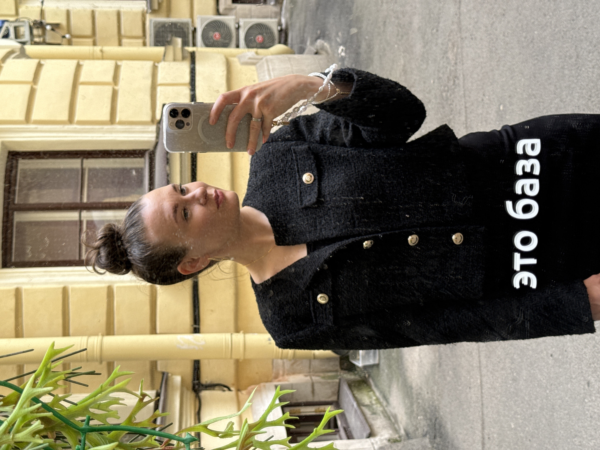The image shows a young Caucasian woman with her hair in a bun, wearing a black tweed jacket with gold buttons and a black top, taking a mirror selfie. She is holding a silver iPhone in her right hand. She is also wearing jewelry, including a thin gold necklace, a ring on her right ring finger, and a multi-strand pearl bracelet on her left wrist. The background is a yellow building with architectural details, including decorative elements around a window and pipes. Two air conditioning units are visible on the building's facade. In the foreground, a plant with green leaves is visible on the left side of the image. The text "ЭТО БАЗА" is superimposed in white on the lower part of the image. This text is in Cyrillic and translates to "THIS IS BASE". The scene is likely in a courtyard or alleyway in Saint Petersburg, Russia, given the architectural style of the building and the presence of Cyrillic text.