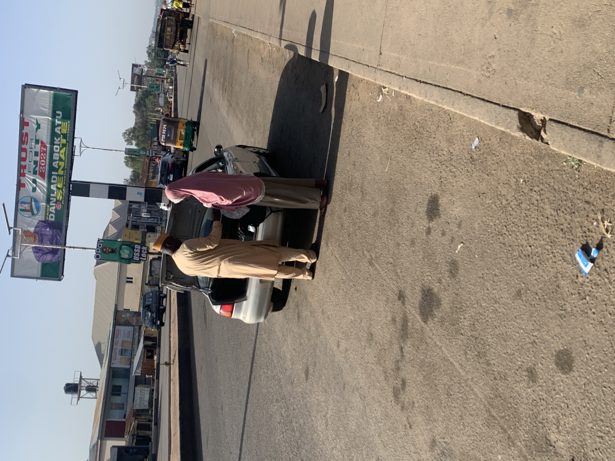 The image depicts a street scene in Gura Top, Nigeria. Two men are standing near a parked silver car on the side of the road. One man is wearing a tan robe and a small hat. The other is wearing a pink robe and pants. In the background, there is a political billboard advertising PROF DANLADI ABOK ATU for SENATE, a billboard advertising Opay, various buildings, street signs, and other vehicles, including a yellow and green tuk-tuk. The road is paved and shows signs of wear and tear. There is debris along the curb. The sky is clear and blue. The car's license plate partially reads "PTA-9894."