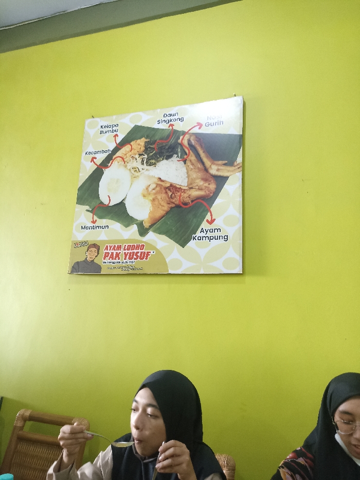 The image captures an indoor scene, likely a restaurant or eatery in Tulungagung, Indonesia, featuring two women having a meal. A bright lime green wall serves as the background, adorned with a large food advertisement poster.
The woman on the left, wearing a black hijab and a light-colored top, is actively eating, bringing a spoonful of food to her mouth with a focused expression. To her right, another woman, also in a hijab and wearing glasses, is partially visible, with a face mask pulled down below her chin. Part of a woven chair back is visible next to the first woman.
Dominating the upper half of the wall is a poster advertising "AYAM LODHO PAK YUSUF." The poster displays an overhead view of a dish, presumably Ayam Lodho, served on a banana leaf. Various components of the meal are labeled with red arrows: "Nasi Gurih" (Savory Rice), "Daun Singkong" (Cassava Leaves), "Kelapa Bumbu" (Seasoned Coconut), "Kecambah" (Bean Sprouts), "Mentimun" (Cucumber), and "Ayam Kampung" (Free-range Chicken). Below the main title, smaller text reads "asli Tulungagung sejak 1987," indicating its origin in Tulungagung and establishment year. A cartoon illustration of an older man in a traditional cap, likely Pak Yusuf, is also part of the branding. The overall setting suggests a casual dining experience.
