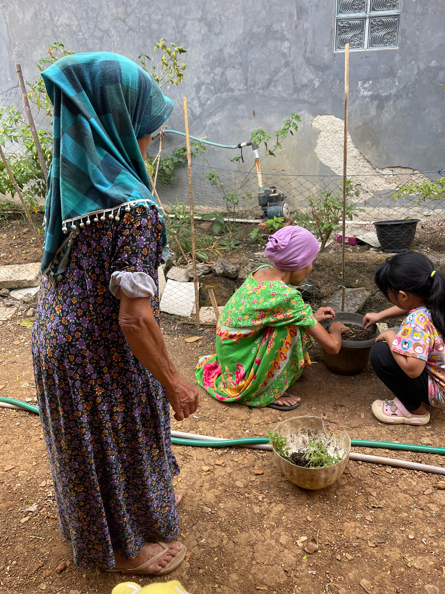 The image depicts an outdoor scene in the city of Pabuaran, Indonesia, where three individuals are engaged in gardening activities. The foreground is dominated by a woman standing, wearing a long, floral-patterned dress and a turquoise headscarf. Her body is oriented to the left, her back to the viewer, as she looks towards two other people. To her right, a younger woman, also wearing a headscarf, this one purple, is crouched down, dressed in a vibrant green, floral-patterned garment. She is working with soil in a large pot. Beside her, a young girl, wearing a colorful t-shirt with cartoon characters and black pants, is also crouched and actively participating in the gardening by handling soil in the same pot. In the middle ground, a metal water pump is visible near a fence and some greenery. The background consists of a textured, grey wall, typical of residential structures in the area. The ground is covered in loose soil and small stones, with a green garden hose running horizontally across the scene. A broken, shallow bowl containing small green plants and soil is placed on the ground in the lower right quadrant.