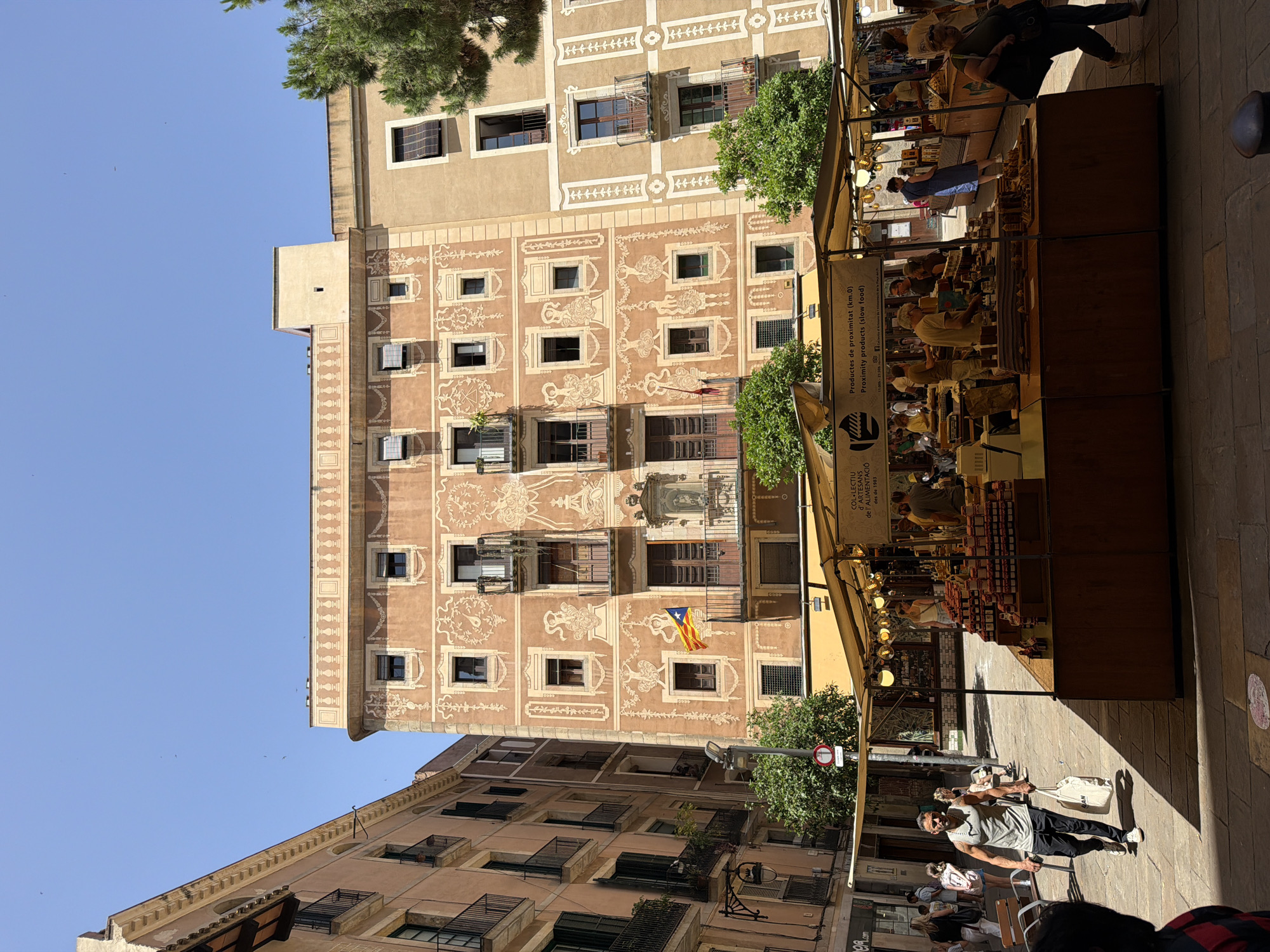 The image depicts a bustling outdoor market in the Gothic Quarter of Barcelona, Spain, under a clear blue sky. In the foreground, a stall is laden with jars of preserves, with several people browsing and interacting with vendors. The stall is covered by a beige awning adorned with decorative lights. To the left, a man walks past carrying a white tote bag, dressed in a sleeveless grey t-shirt and black pants.
Dominating the background is a historic building with distinctive ornate architectural details, painted in warm terracotta hues with contrasting cream decorative friezes featuring stylized figures and floral motifs. Several balconies with black iron railings punctuate the facade. A Catalan flag is visible hanging from one of the balconies. Adjacent to this building is another similar structure, also with balconies and ornate detailing. Lush green trees are interspersed between the buildings and the market stalls, adding a touch of nature to the urban landscape. The ground is paved with traditional cobblestones. The overall scene conveys a vibrant and lively atmosphere of local commerce and historical architecture.