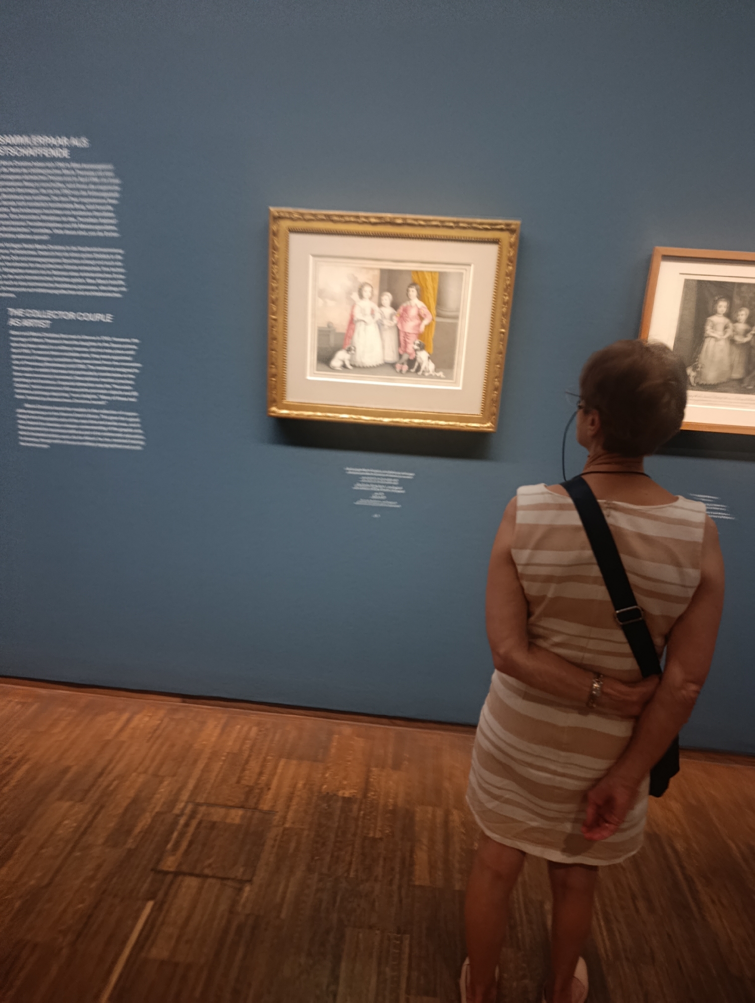 A woman stands in a museum, facing away from the camera, looking at a painting on a blue wall. She is wearing a striped dress and has her arms crossed behind her back. The painting in front of her is framed in gold and depicts two children and a dog. To the right of this painting is another framed artwork, a sketch of two children. Text panels are visible on the wall to the left of the paintings, one with the title "THE COLLECTOR COUPLE AS ARTIST". The museum floor is made of polished wood. The lighting suggests it is daytime.