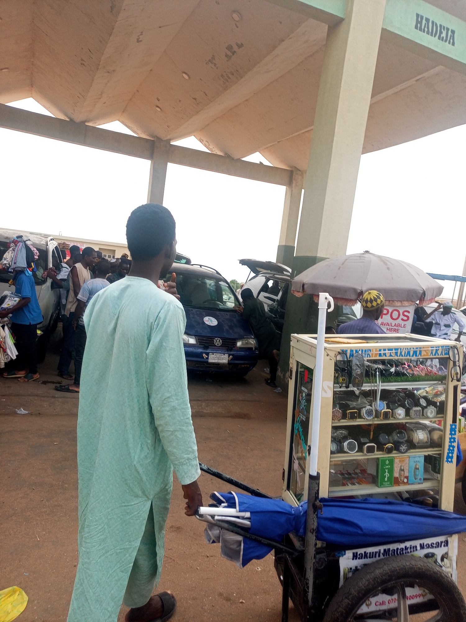 This image captures a bustling outdoor scene, likely a market or a transport park, in what appears to be Gombe, Nigeria. The time of day is daylight, under what seems to be an overcast sky or a large shade structure.
In the foreground, a man, seen from behind, is the central figure. He wears a light green, long-sleeved garment, possibly a thobe or kaftan, and dark sandals. He is pushing a cart that serves as a mobile display stand. The top part of the cart is a glass-encased shelf displaying various items, including watches, sunglasses, and small bottles, possibly perfumes. Folded blue umbrellas are stored below the display case. Text on the side of the cart reads "Hakuri Matakin Nasara" and a partial phone number starting "Call: 070".
In the midground, several people are gathered around vehicles, specifically two vans or minibuses. One dark blue van has its rear door open, and people are either loading or unloading goods. Another white van has items, possibly clothing, stacked on its roof. The atmosphere suggests commercial activity and travel.
The background features a large, concrete architectural structure, likely a roof or shelter for the area. On one of the beams, the word "HADEJA" is painted in dark letters. Beneath this structure, further back, another vendor stall is visible, featuring a large grey umbrella and a sign that clearly states "POS AVAILABLE HERE," indicating the presence of point-of-sale services. People are scattered throughout the scene, engaging in various interactions. The overall impression is one of a busy, functional public space.