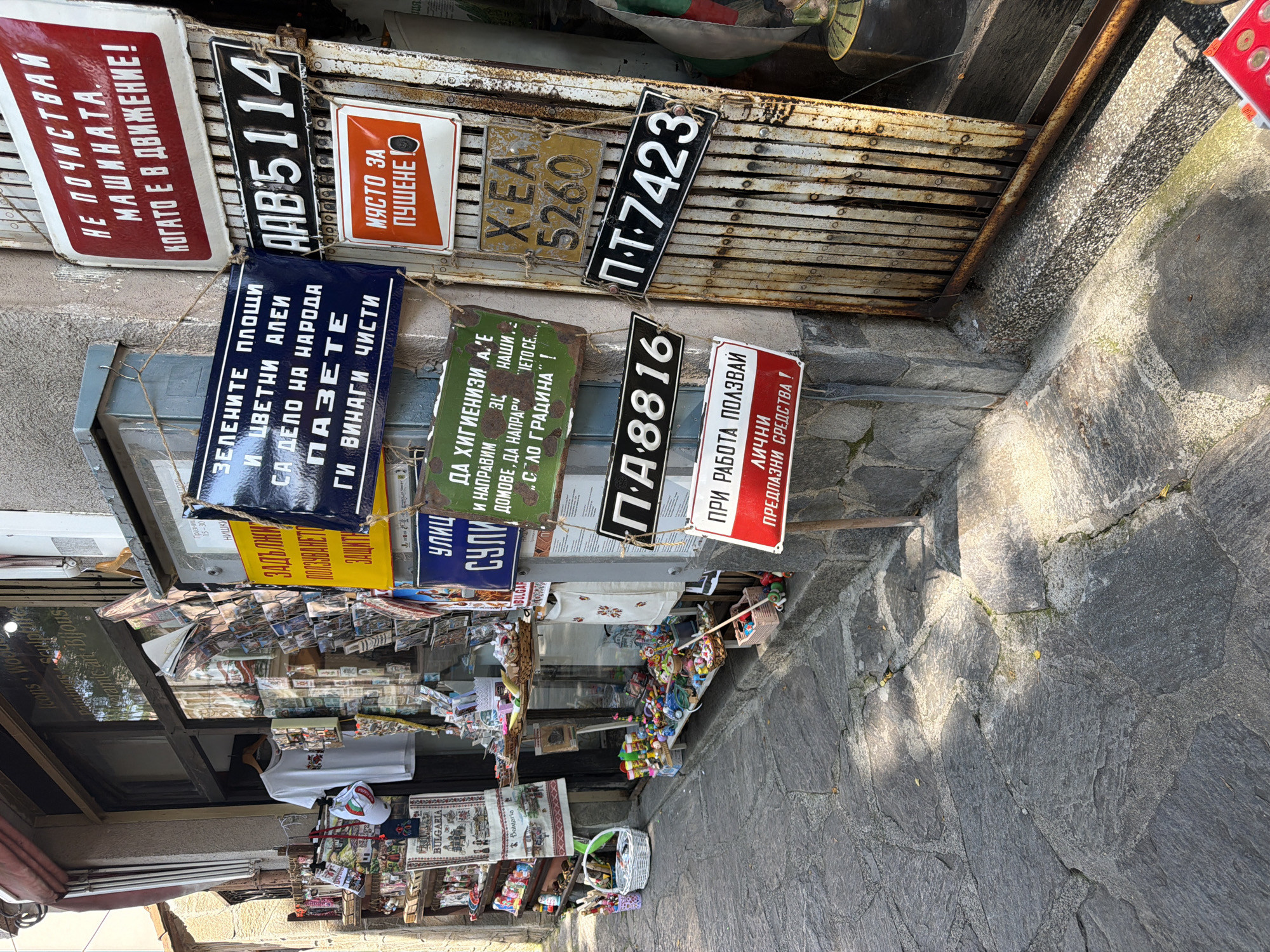 The image depicts a detailed view of a shop exterior and an adjacent building facade on a sunlit street in Plovdiv, Bulgaria. The foreground features a stone-paved sidewalk, showing natural wear and scattered small yellow debris, with distinct patterns of sunlight and shadow. On the left, a shop window displays various items, primarily souvenirs and handcrafted goods. Visible elements include a white t-shirt on a hanger, a white baseball cap with embroidery, shelves stocked with small trinkets, textiles featuring "BULGARIA" branding, and a rotating stand with postcards or books. Reflections in the window show partial text like "Icons • Wood" and "Paintings • Sculpture." Below the window, along the sidewalk, are several baskets filled with colorful small objects, suggesting items for sale. A red awning partially covers the top left of the shop entrance. To the right, a weathered, rusty, accordion-style metal security gate is partially retracted, revealing a dark interior. The gate and the adjacent building wall are heavily adorned with numerous vintage-style metal signs, mostly rectangular and displaying Cyrillic text in various colors (red, blue, green, black, orange, yellow, white). These signs appear to be old warning or informational plaques and several repurposed license plates. Specific visible signs include: * A red and white sign at the top right: "НЕ ПОЧИСТВАЙ МАШИНАТА, КОГАТО Е В ДВИЖЕНИЕ!" (DO NOT CLEAN THE MACHINE WHEN IT IS IN MOTION!) * A dark blue and white sign below it: "ЗЕЛЕНИТЕ ПЛОЩИ И ЦВЕТНИ АЛЕИ СА ДЕЛО НА НАРОДА ПАЗЕТЕ ГИ ВИНАГИ ЧИСТИ" (GREEN AREAS AND FLOWER ALLEYS ARE THE WORK OF THE PEOPLE. KEEP THEM ALWAYS CLEAN) * A black and white sign with characters "AAB 5114" (resembling a license plate). * An orange and white sign with a cigarette icon: "МЯСТО ЗА ПУШЕНЕ" (SMOKING AREA). * A partially visible green and white sign with text related to hygiene and "CITY GARDEN." * Three black and white or yellow and black signs, clearly old license plates, displaying "X•EA 5260", "П.A.8816", and "П.Т-7423". * A red and white sign at the bottom left: "ПРИ РАБОТА ПОЛЗВАЙ ЛИЧНИ ПРЕДПАЗНИ СРЕДСТВА!" (WHEN WORKING USE PERSONAL PROTECTIVE EQUIPMENT!). Leaning against the wall near the bottom right corner of the image, partially obscured by the metal gate, is a Bulgarian national flag. On the ground at the far bottom right, a small red tray or box contains several coins or tokens. No people are visible in the