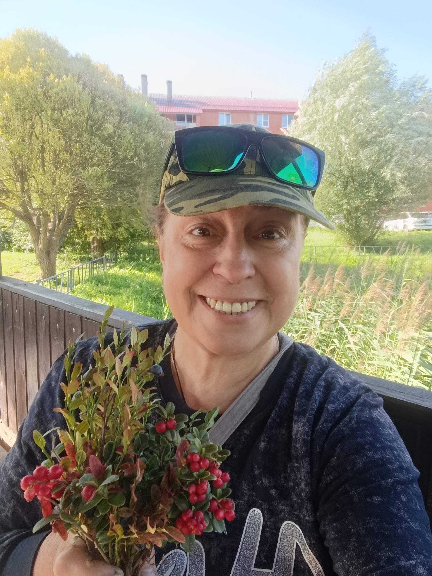 A woman with a camouflage baseball cap and sunglasses on her head smiles at the camera while holding a bouquet of lingonberries and blueberries. She is standing on a wooden porch, with a railing to her left. In the background, there are trees, a building, and tall grass. The scene appears to be in a rural or suburban setting, possibly in the city of Vereya, Russia. The lighting suggests it is daytime, with bright sunlight casting shadows.