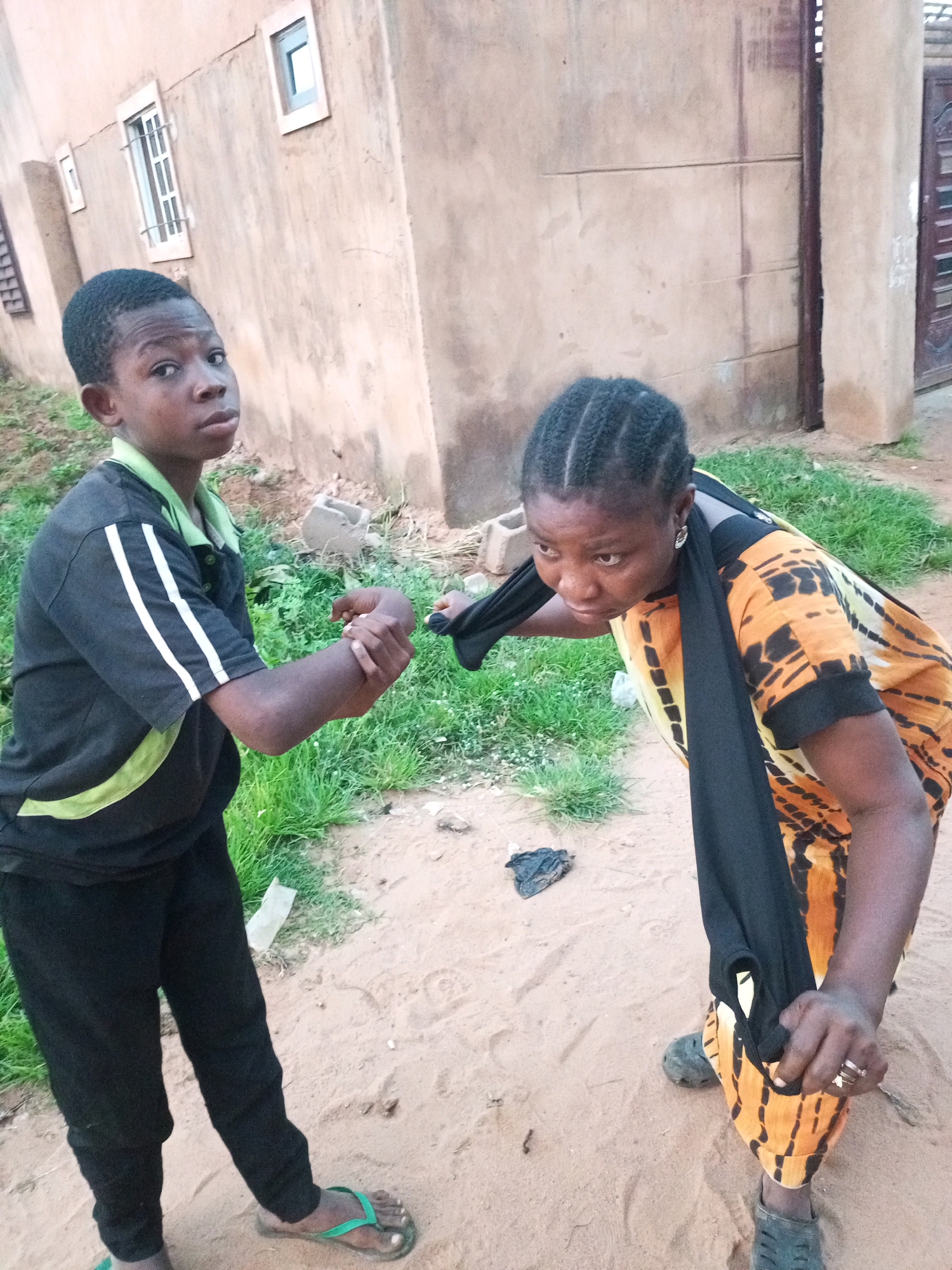 In a sandy, outdoor setting in Tempure, Nigeria, a young boy wearing a black polo shirt with lime green accents and black pants, and a woman wearing an orange and black patterned dress are interacting. The boy is holding the woman's right wrist with his left hand. The woman is pulling a black garment, possibly a scarf or shirt, with her right hand. She is also wearing dark shoes. The background consists of a building with a textured, light brown wall and some greenery. The ground is sandy with sparse grass.
