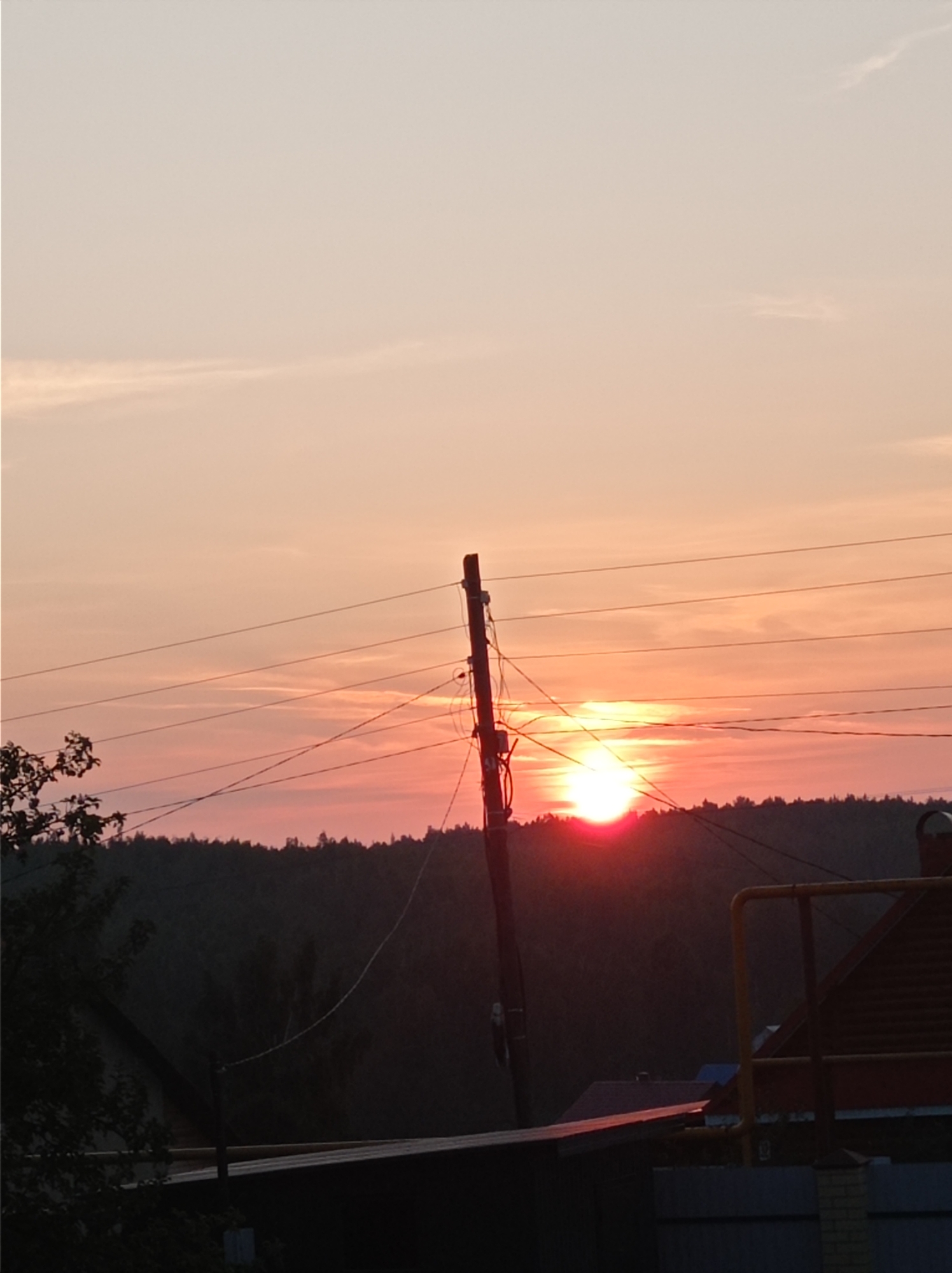The image depicts a sunset over a wooded area on the outskirts of Degtyarsk, Russia. A utility pole with multiple wires runs through the frame, silhouetted against the vibrant sky. The sun, a bright orb of red and orange, is partially obscured by the horizon line of dark trees. In the foreground, the rooftops of several houses are visible, along with some fencing and vegetation. The scene is quiet and appears to be taken from a residential area looking towards a natural landscape.