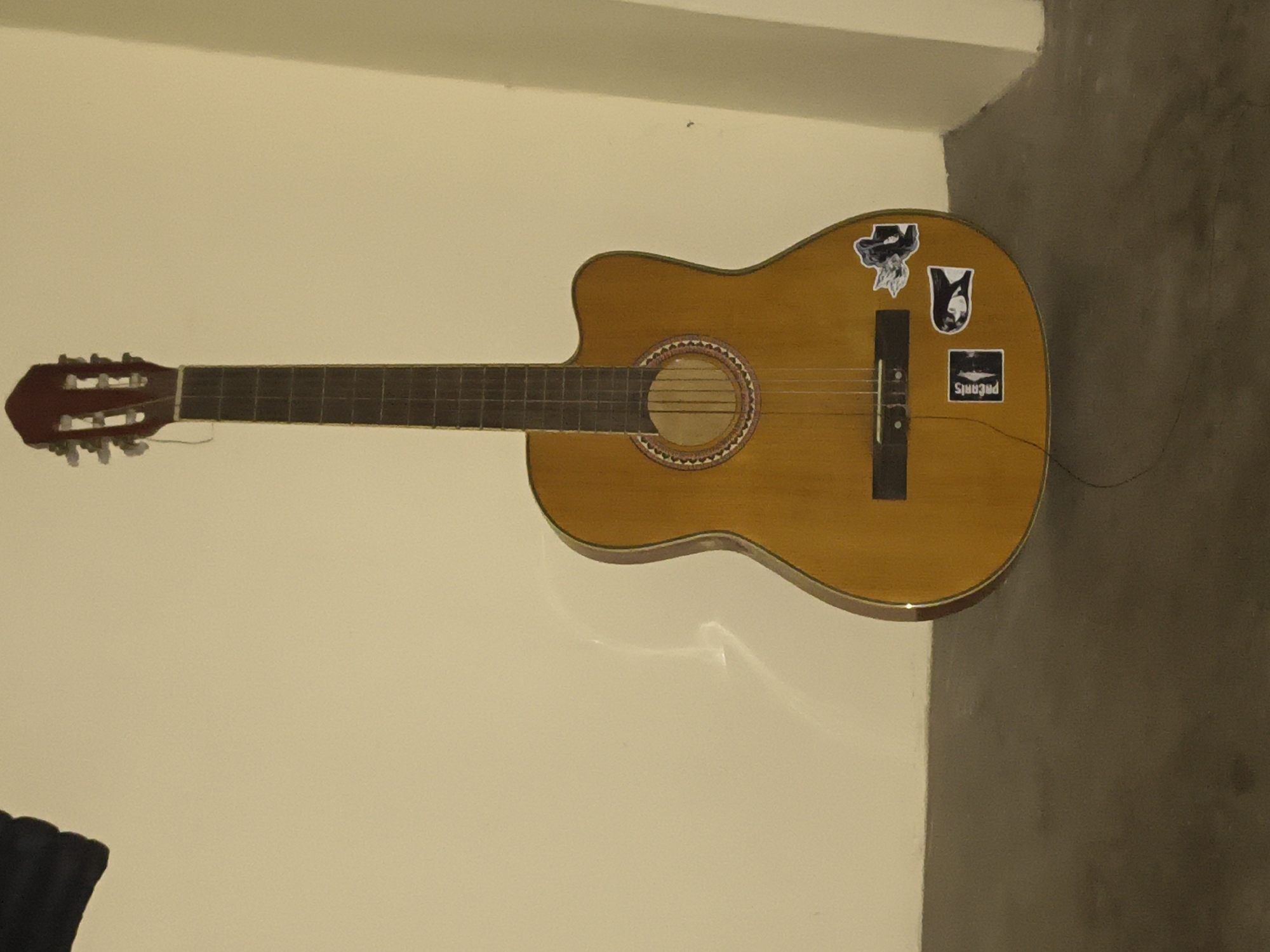 An acoustic guitar with a cutaway body and a natural wood finish stands upright against a plain, light-colored wall. The guitar has six strings, a dark fretboard with inlaid fret markers, and a rosette around the soundhole. Several stickers are adhered to the body of the guitar, including one that appears to be a black and white portrait and another with text that is partially obscured. The guitar is positioned in the foreground, with its base resting on a dark gray floor. The background consists of the wall and a corner where two walls meet. There is a shadow cast on the wall behind the guitar, suggesting a light source from the left. The scene is illuminated with soft lighting, creating a somewhat muted atmosphere. The location is not specified.